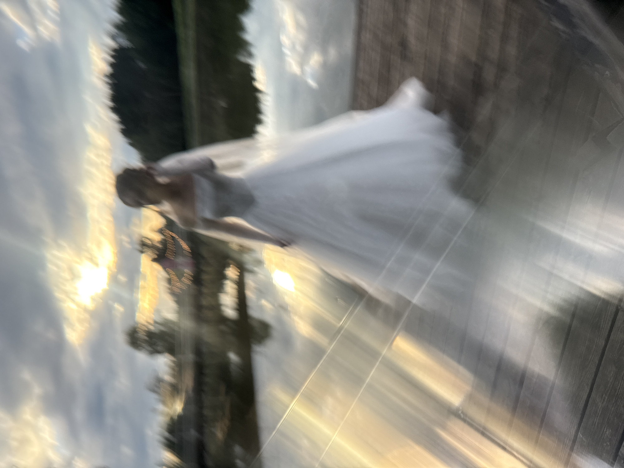 An out-of-focus, full-shot, vertical, medium-wide photograph captures a person in a white wedding dress standing on a wooden dock next to a body of water. The person is positioned in the center of the frame, facing to the right, with their head slightly turned towards the viewer. The wedding dress is a ball gown with an off-the-shoulder neckline. A veil extends from the back of the head. The background consists of a cloudy sky with the sun visible and casting bright rays, trees along the far bank of the water, and a few indistinct structures. The lighting is characteristic of sunset or sunrise, with strong golden hues. The wooden dock occupies the foreground, with the water reflecting the sky and light. The overall image is characterized by significant motion blur, creating a streaked effect across the scene, especially in the water and on the dock.