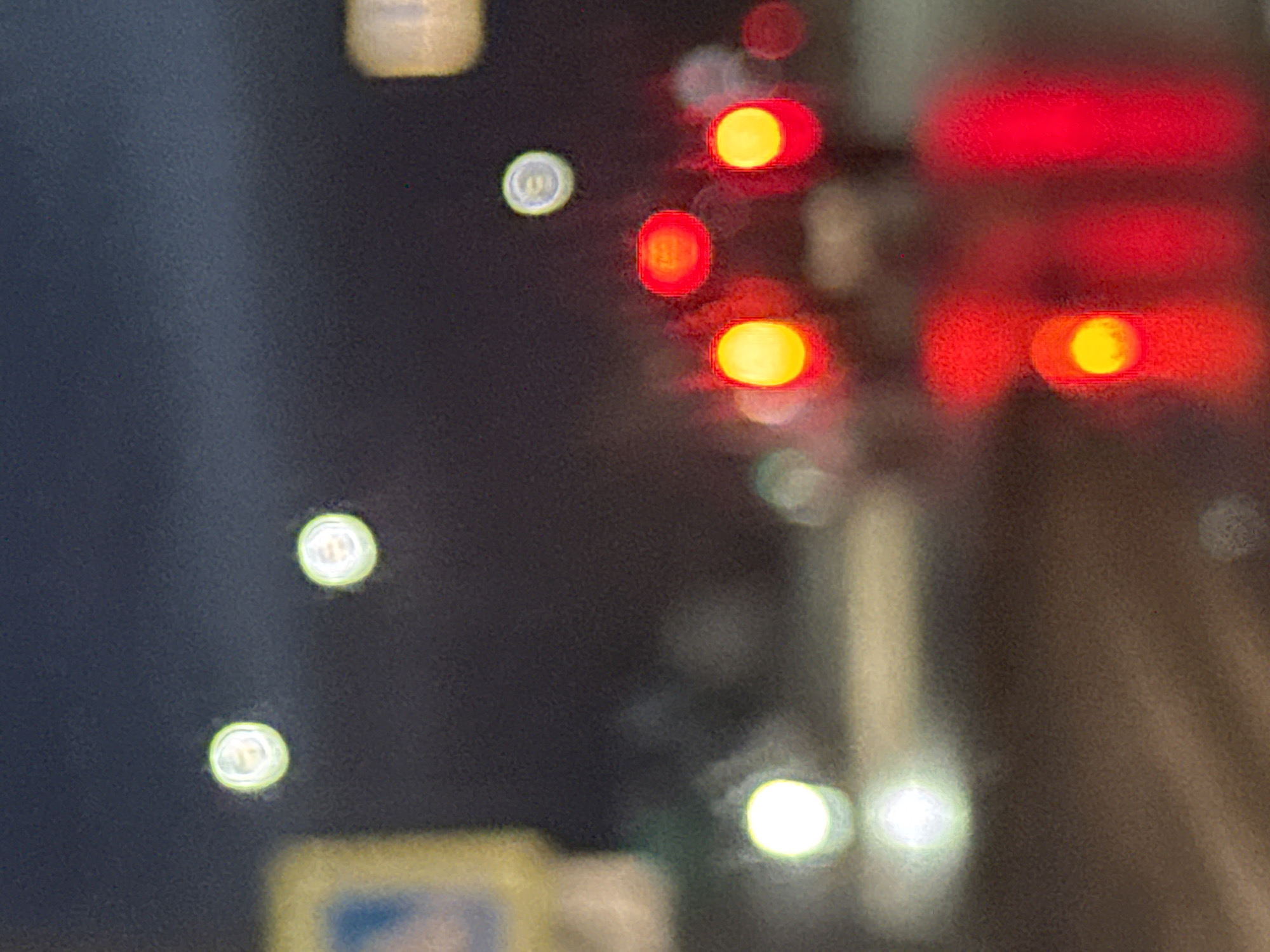 The image appears to be an out-of-focus photograph of traffic lights and headlights at night. The red lights of the brake lights are visible and the headlights are out of focus. The location is not discernible from the information in the image.