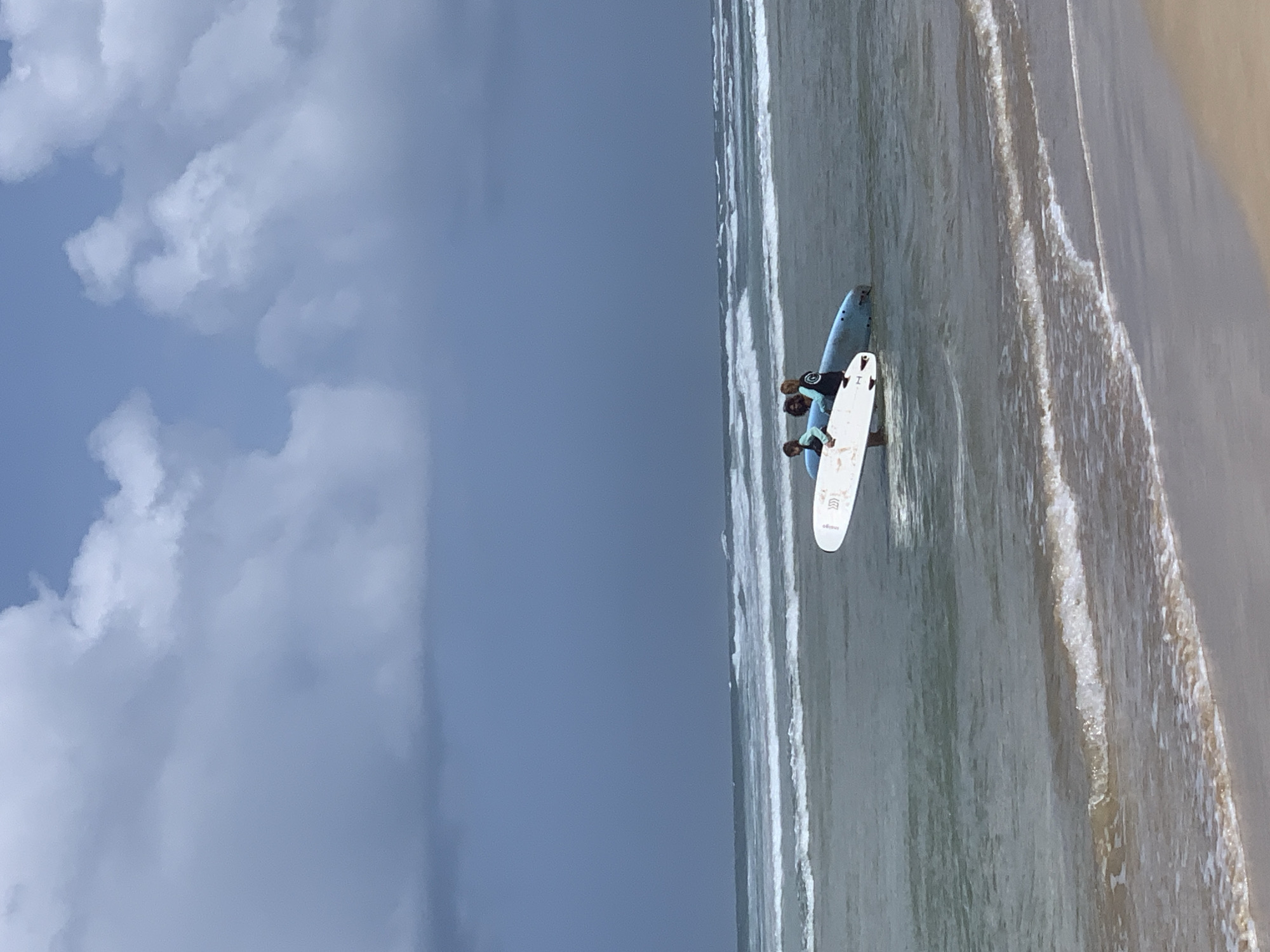 In Kathaluwa, Sri Lanka, two individuals are in the shallow ocean, each carrying a surfboard. The person on the left holds a white surfboard, while the person on the right is positioned behind a blue surfboard. Both are facing towards the ocean. The water is calm, with gentle waves lapping at the sandy shore in the foreground. The sky above is partly cloudy, with a mix of darker and lighter clouds. The scene appears to be set on a beach.