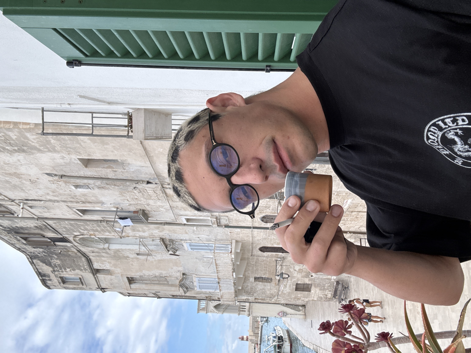 The image captures a close-up of a person on a balcony, observing a historic coastal town below. The subject is a young individual with short, light-colored hair, possibly bleached or patterned, and is wearing round, dark sunglasses. They are dressed in a black t-shirt featuring a partially visible white circular logo on the right side of the chest; text like "OLD LTD" and "201" are discernible within the logo, alongside a stylized creature. The person holds a lit cigarette in their right hand and a small, two-toned cup or container (with a terracotta base and grey top) in their left. Their facial expression is neutral.
In the foreground, green louvered shutters and a white balcony ledge are visible at the top, along with red succulent-like plants and green spiky plants in pots on the right. The background reveals a picturesque townscape, consistent with Monopoli, Italy, under a bright, partly cloudy sky. Weathered, light-colored stone buildings with numerous windows and balconies line a waterfront. Some windows have blue louvered shutters, and laundry is seen hanging to dry on one building. Below, a canal or harbor features at least one boat and a small lighthouse. Two small figures are visible relaxing on a lower level near the water.
The scene depicts a sunny, clear day, likely midday or early afternoon, with the person enjoying a moment of relaxation and observation from their elevated position.