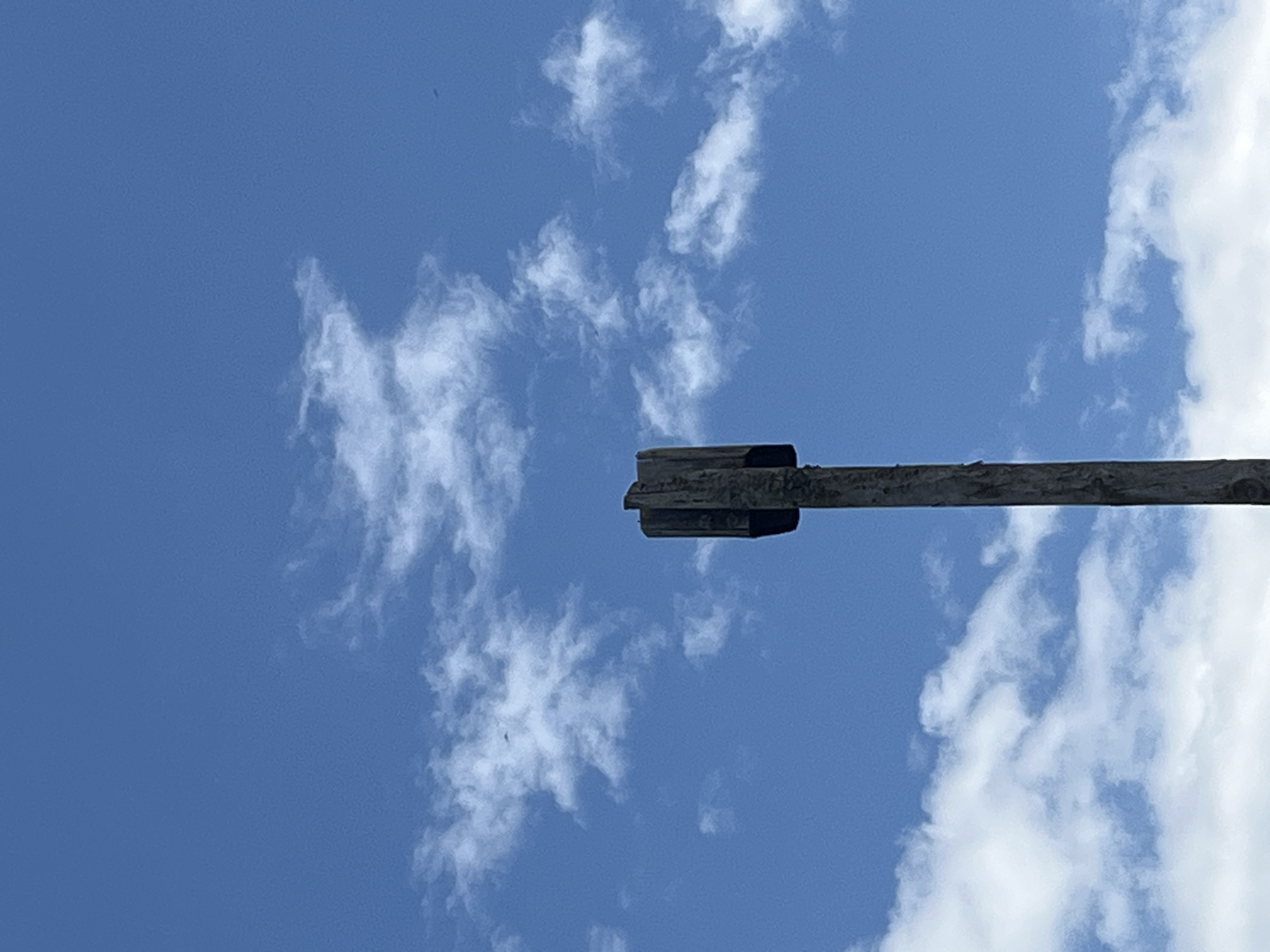 A close-up, low-angle shot shows a weathered wooden post reaching upwards into a bright blue sky filled with wispy white clouds. The top of the post is capped with four vertical wooden planks, forming a square, open-ended structure. The post is centrally located in the frame, with the sky dominating the upper portion and the bottom edge of the post visible. The overall lighting suggests daylight. This scene is unlikely to be in Pamyati 13 Bortsov, Russia, as the image lacks any contextual urban or geographical features.