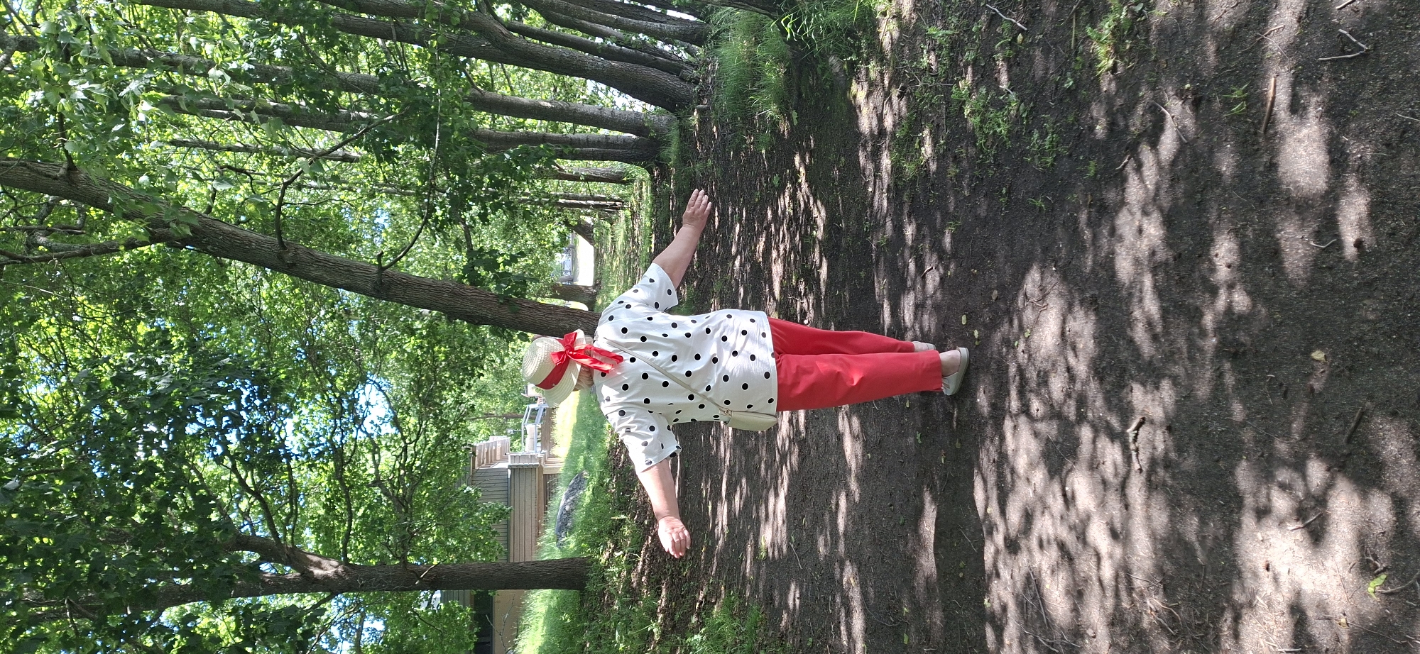 The image shows a person from behind walking on a dirt path through a wooded area. The person is wearing a white shirt with black polka dots, red pants, a straw hat with a red ribbon, and light-colored shoes. They have their arms extended outwards as if enjoying the surroundings. A small, light-colored bag is visible on their left side, possibly a purse or cross-body bag. The path is dappled with sunlight filtering through the dense green foliage of trees lining the right side of the path. On the left side of the path, there is a grassy embankment with some trees and a glimpse of a wooden building in the background. The overall environment suggests a park or nature trail. The presence of a building in the background may indicate proximity to a park amenity or residential area within Lahti, Finland. The lighting suggests it is daytime, likely late morning or early afternoon due to the bright sunlight and distinct shadows.