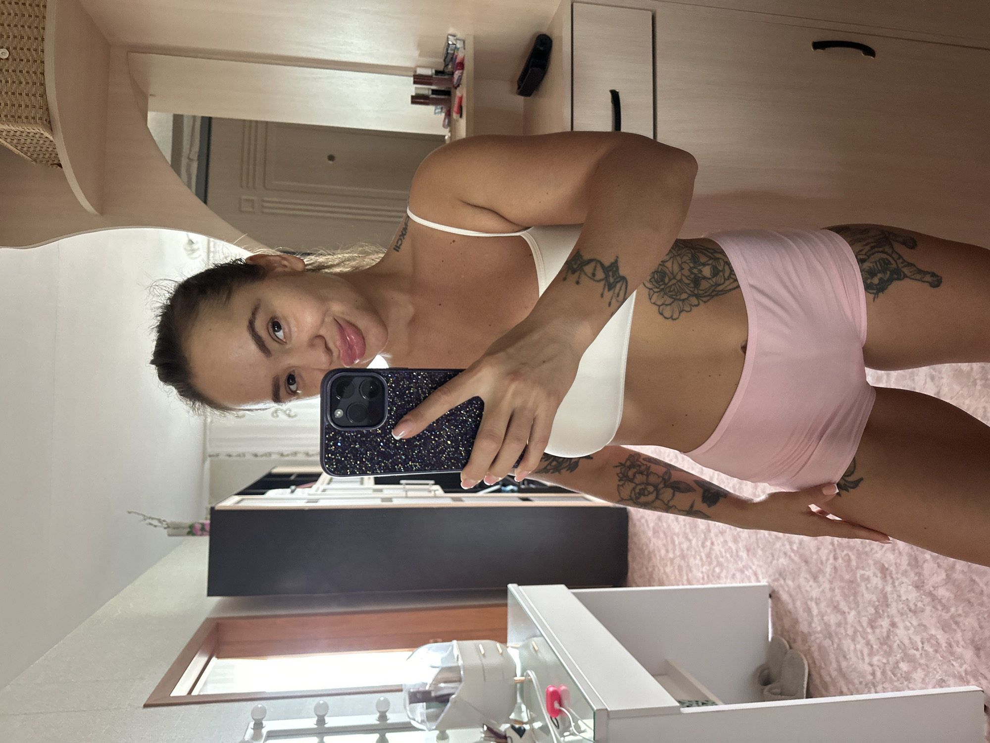 A woman with dark hair pulled back in a bun and fair skin is taking a selfie in a mirror. She is wearing a white crop top and pink shorts. She has multiple tattoos on her arms and torso, including a lion on her hip and a tiger on her thigh. She is holding a smartphone with a sparkly dark case in her right hand. In the background, there is a white vanity with makeup, a dark closet, and a beige dresser with a mirror. The floor is covered in a pink rug. The scene appears to be a bedroom or dressing area in a residential setting. There is no indication of the specific location within Yuzhnouralsk, Russia.