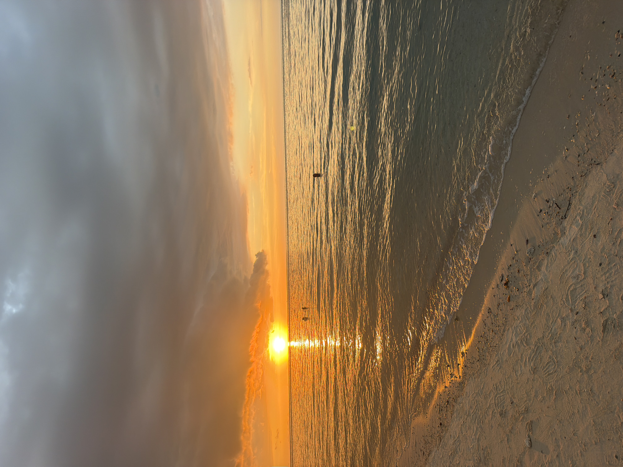 The image depicts a serene sunset scene at a beach in Baan Tai, Thailand. The sky is a gradient of colors, with dark gray clouds at the top transitioning to vibrant orange and yellow hues near the horizon. A brilliant sun is setting over the calm sea, casting a shimmering reflection on the water's surface. The water itself is a blend of deep blue and golden tones. On the sandy shore in the foreground, there are faint tracks, possibly from beachgoers or sea creatures. A few individuals are visible in the water, appearing to be swimming or wading. One person is further out in the sea, appearing as a dark silhouette. The overall atmosphere is tranquil and picturesque.