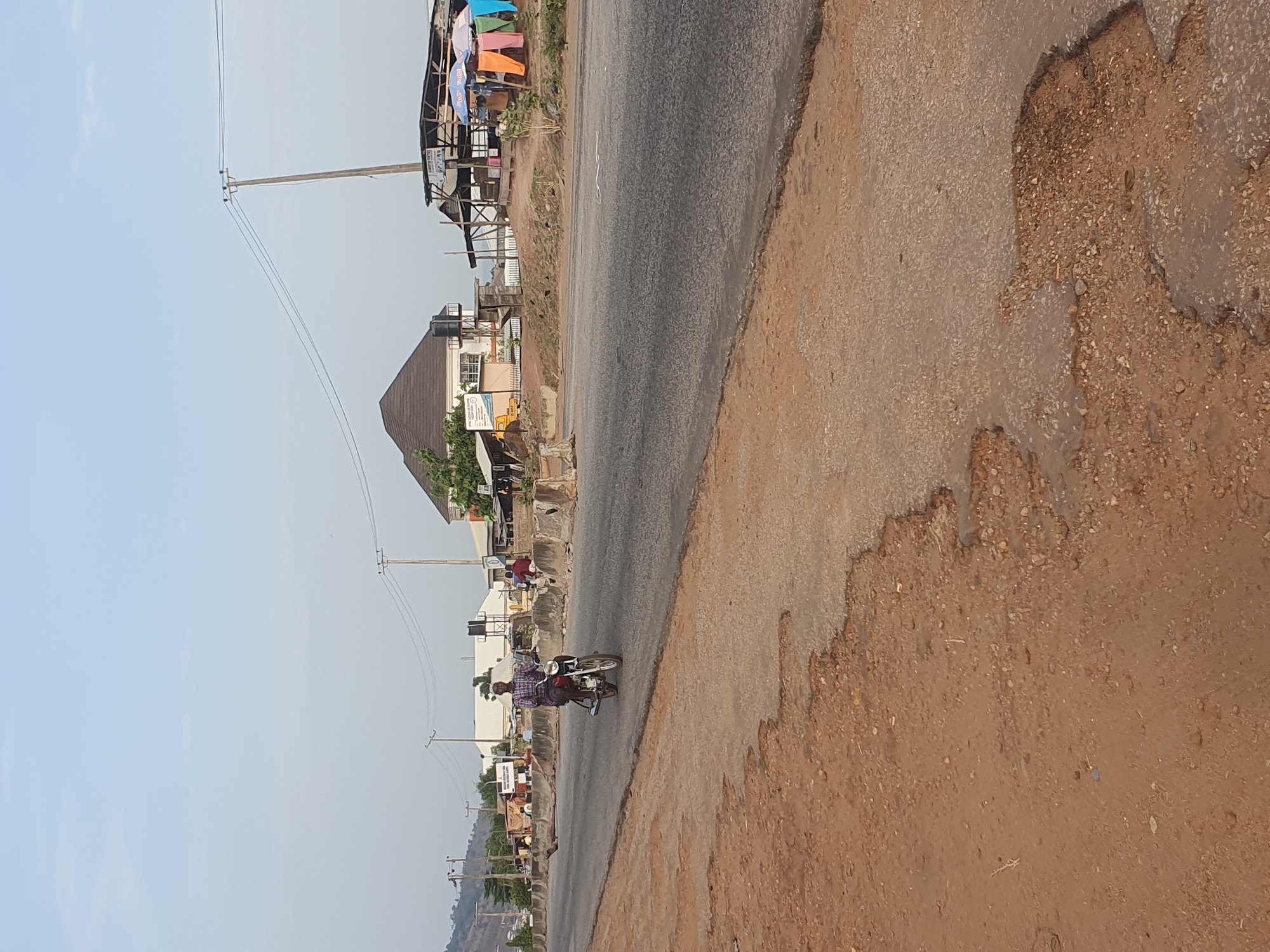The image shows a street scene in Lokoja, Nigeria. A motorcycle with two riders is traveling on the paved road. The road is bordered by a wide, unpaved shoulder. Buildings are visible along the roadside, including shops and houses. Power lines run parallel to the road. The sky is clear and pale. The road surface shows signs of wear and tear.