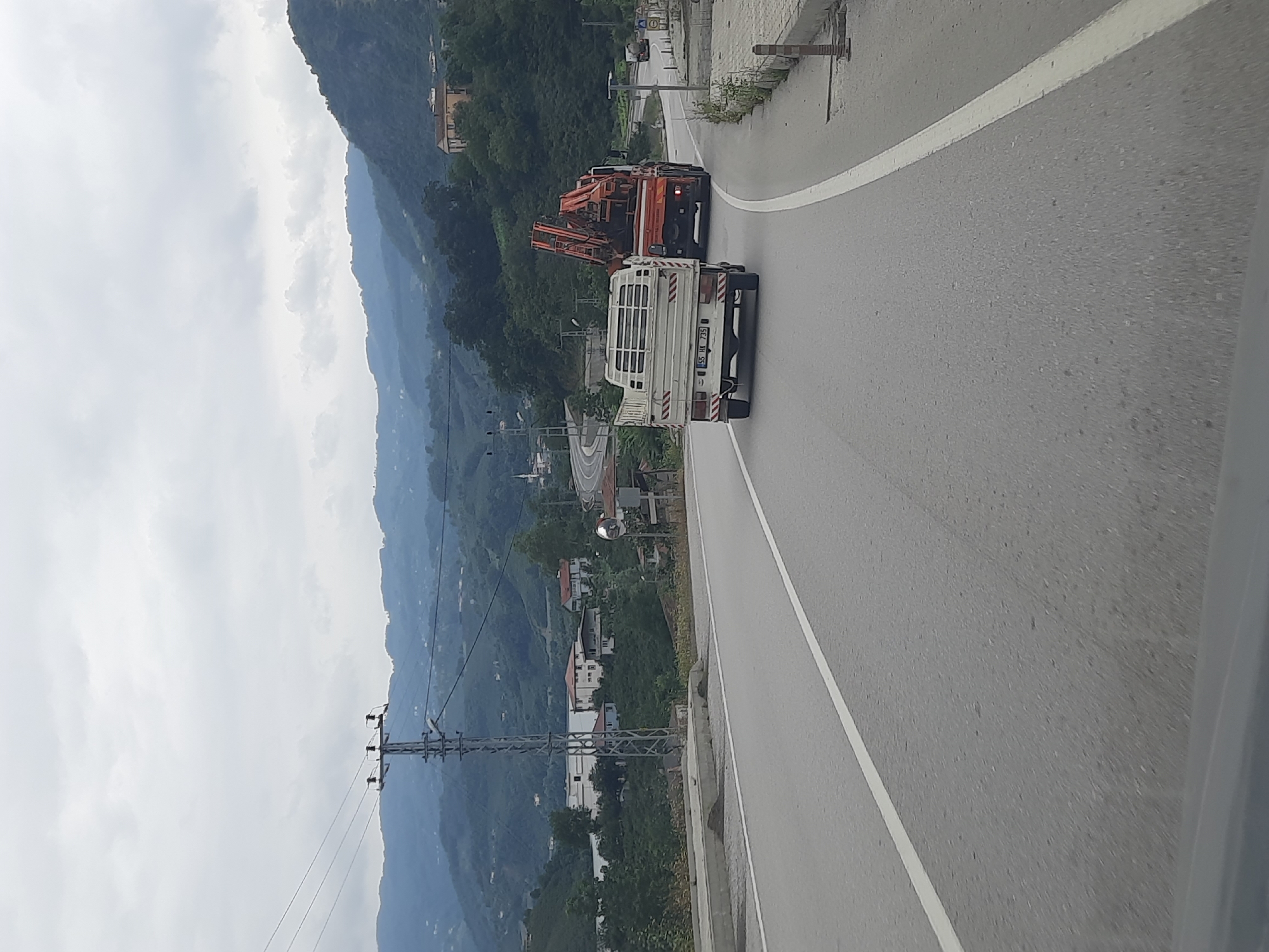The image depicts a grey, overcast day with a mountainous, vegetated landscape. A paved road with a white lane marker curves through the scene. In the foreground, a white pickup truck with a flatbed is driving on the road. Behind it, a large orange truck with a crane attachment is also in motion. In the background, several buildings are visible nestled into the hillsides, along with a tall electrical pylon. The location is identified as being in Ayvacık, Turkey.