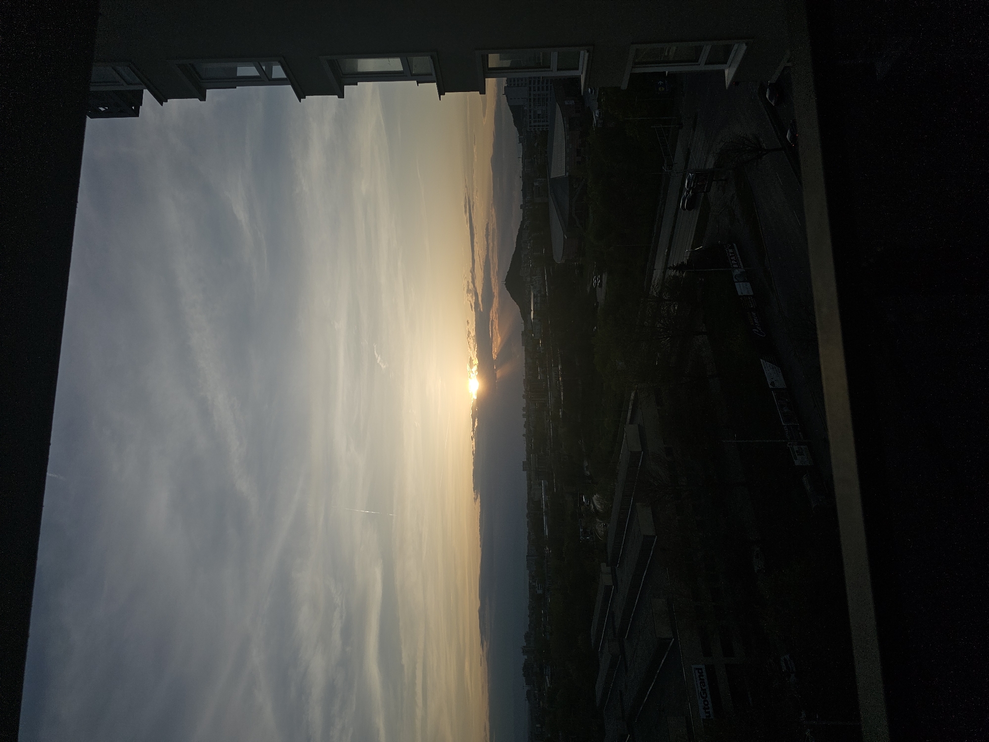 The image presents a sunset view, partially framed by a building's window. The sky transitions from a pale blue with streaky clouds to a golden hue near the horizon, where the sun is setting. Below the horizon, a silhouetted cityscape stretches into the distance, possibly Plovdiv, Bulgaria. The skyline is punctuated by buildings and what may be a distant hill or mountain. A street is visible to the right, with some indication of vehicles and possibly pedestrian structures. The immediate foreground is dominated by the building structure framing the view, and is dark.