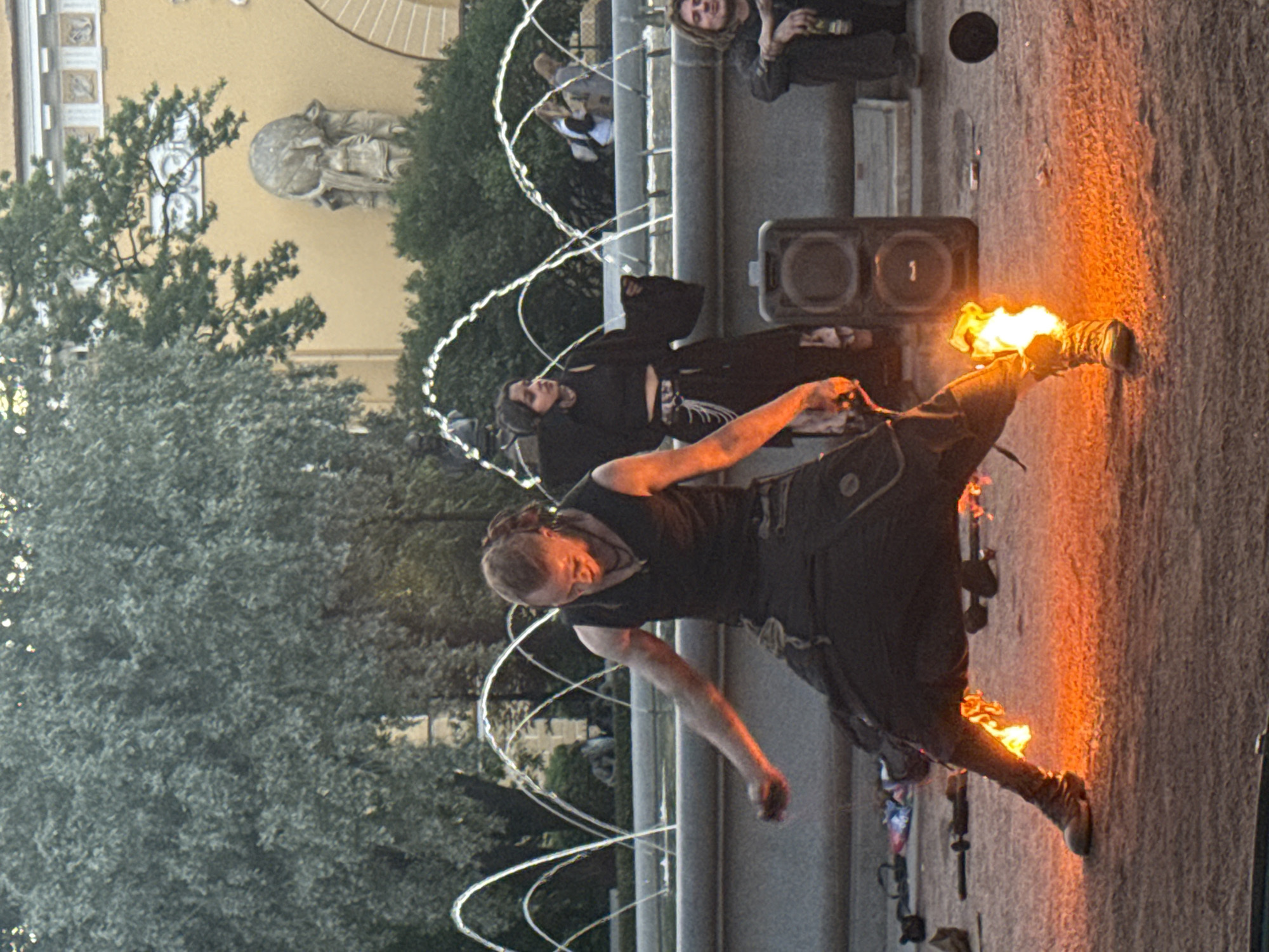 The image captures a street performance in Saint Petersburg, Russia, likely in an outdoor public square or park. In the foreground, a male performer with dreadlocks and dark clothing is engaged in fire dancing. Flames are visible around his legs and feet, suggesting he is performing with fire-attired props. He is looking down, focused on his actions, with his arms extended. To his right, a female performer in dark, loose-fitting clothing, including a crop top and wide pants, stands with her hands outstretched, observing the scene. Behind them, a large speaker is placed on the ground, indicating the use of amplified music for the performance. Several water jets from a fountain arc in the background, adding to the dynamic atmosphere of the scene. Further in the background, onlookers are visible, as well as architectural elements of a building, possibly a museum or historical structure, with ornate details and statues. The ground appears to be paved with a reddish-brown material, possibly sand or gravel, common in outdoor public spaces. The lighting suggests it is either late afternoon or early evening.