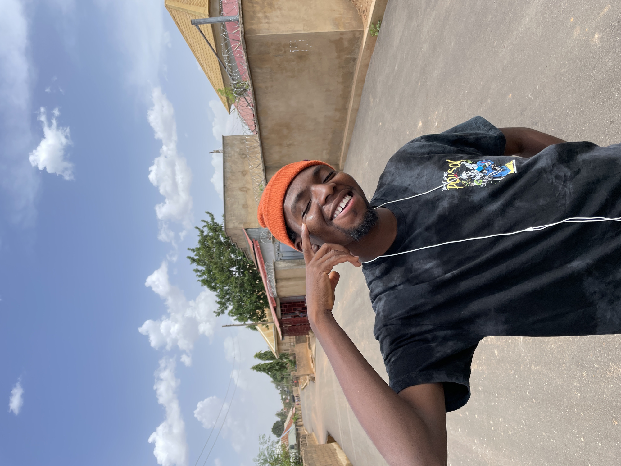 A young man is positioned on a paved street in Gura Top, Nigeria. He is wearing an orange beanie and a dark tie-dye t-shirt with a graphic design. He has dark hair and a beard. He is smiling with his eyes closed, and his right index finger touches his right eye. White earbud wires are visible around his neck. The background includes buildings with barbed wire fencing and a blue sky with scattered clouds.
