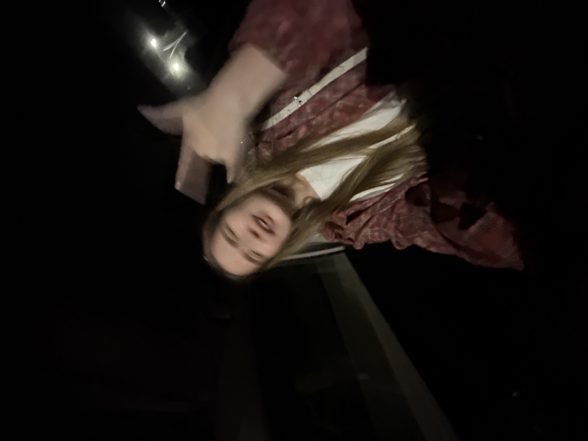 The image shows a young woman holding a cell phone and possibly taking a selfie in Boreti, Montenegro. The photo is taken from a low angle, looking up at the woman. She has long blond hair and is wearing a red and white patterned shirt. It appears to be night, or a poorly lit area. In the background, there are hints of urban features.