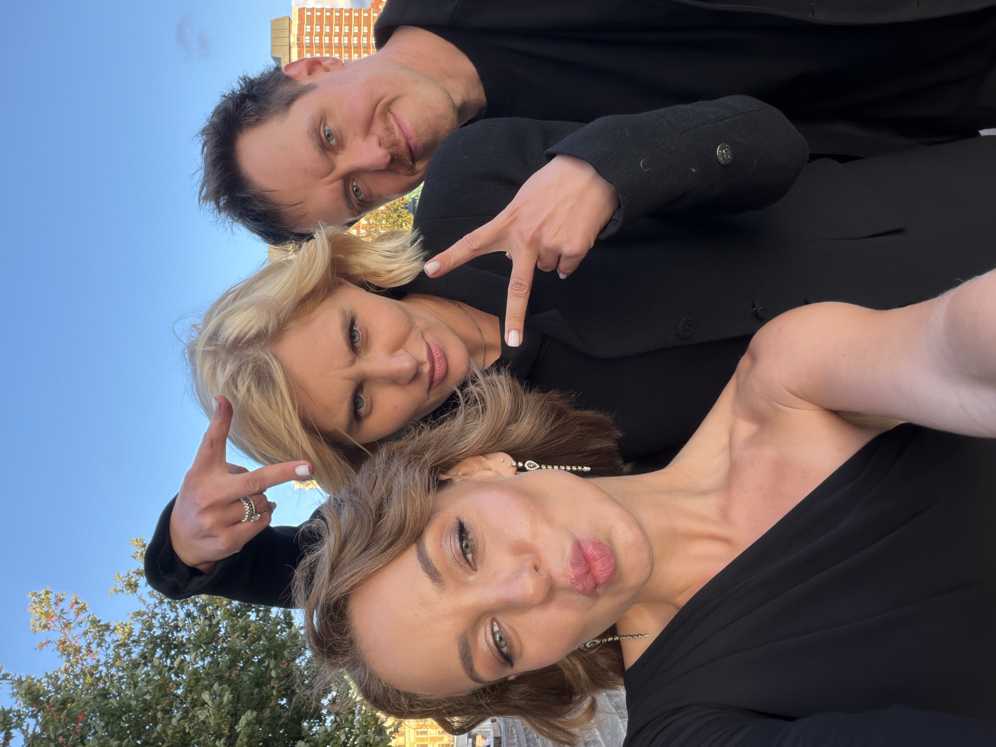 Three individuals, two women and one man, are posing for a selfie outdoors. The woman in the foreground on the left is looking at the camera with her mouth pursed, wearing a black one-shoulder dress and dangling earrings. The woman in the middle has blonde hair and is making a peace sign with her right hand, wearing a black coat. The man on the right has dark hair and a mustache, wearing a black jacket, and is also making a peace sign with his right hand. They are positioned in front of a blurred background of buildings and trees under a clear blue sky. The scene appears to be a casual outdoor gathering. Based on the architecture visible in the background, it is highly probable the location is in Moscow, Russia.