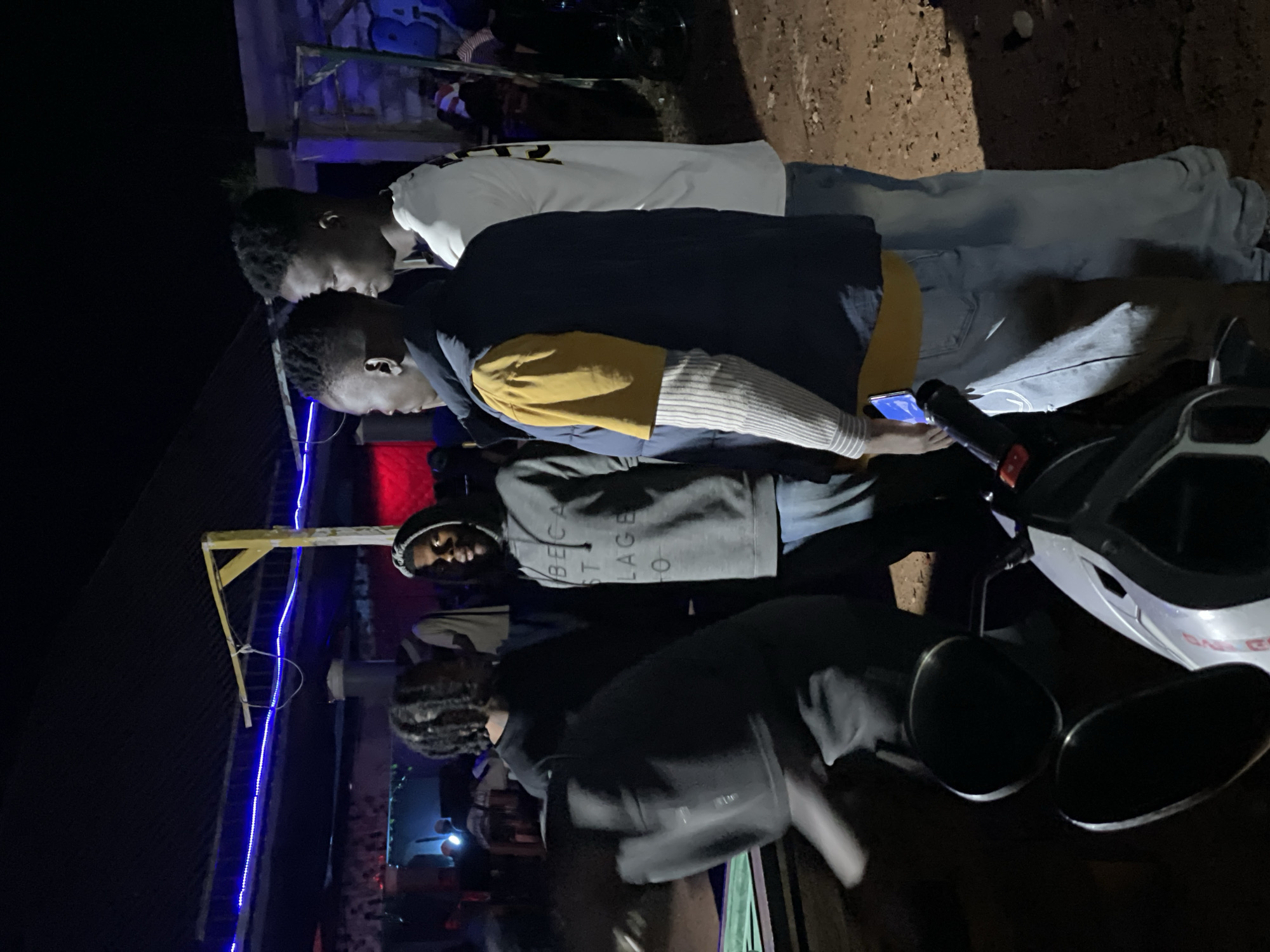 In Durumi, Nigeria, at night, a group of young men are gathered outdoors. The scene appears to be near a structure with a dark roof illuminated by blue string lights. One man in the foreground, wearing a grey hooded sweatshirt with text that reads "BECA ST LAGE O" and blue jeans, faces the camera. To his left, another man with dreadlocks, dressed in a black sweatshirt, is mostly obscured by a black motorcycle in the foreground. To the right of the man in the grey hoodie, two other young men stand side-by-side. One, in a dark vest over a yellow shirt and striped long-sleeved shirt, faces left. The other, wearing a white t-shirt, faces right and appears to be looking away from the camera. The ground is a dirt surface. The lighting is dim, with artificial lights providing illumination.