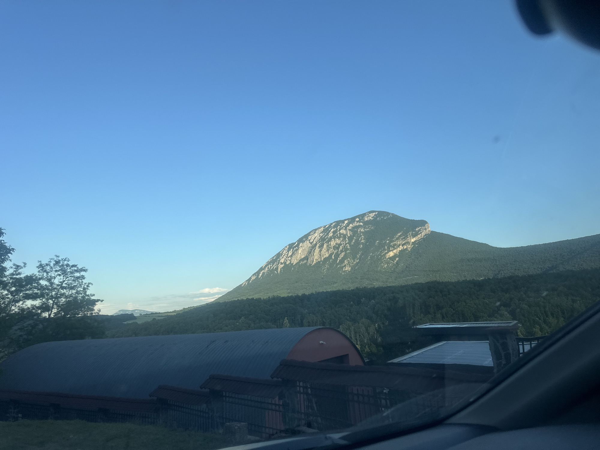 The image displays a serene landscape primarily featuring a majestic mountain under a clear, bright blue sky. The mountain, characterized by a prominent, rocky peak and extensive dark green forested slopes, dominates the upper and mid-portions of the scene.
In the foreground and midground, several man-made structures are visible. A large, dark-colored building with a corrugated, rounded metal roof and a reddish entrance is centrally located. In front of this, a smaller structure with a dark, possibly tiled roof and fence-like elements is partially obscured. To the right, another building features a flat roof, possibly equipped with solar panels, and has visible stone elements on its side. Parts of a vehicle's interior, specifically the dashboard and windshield frame, are seen in the bottom right corner, indicating the photograph was taken from inside a car or similar transport.
Trees with lush foliage are visible on the left, and distant, hazy hills can be discerned on the horizon in the left-midground. The clear sky and soft, directional light suggest either early morning or late afternoon. The weather is fair with no visible clouds. A small, dark smudge, likely a dust particle on the camera lens or sensor, is faintly visible in the sky in some crops.
No people or visible text are present. The scene depicts a rural or semi-rural setting, likely in the Glavatsi region of Bulgaria, showcasing a blend of natural landscape and functional human structures.