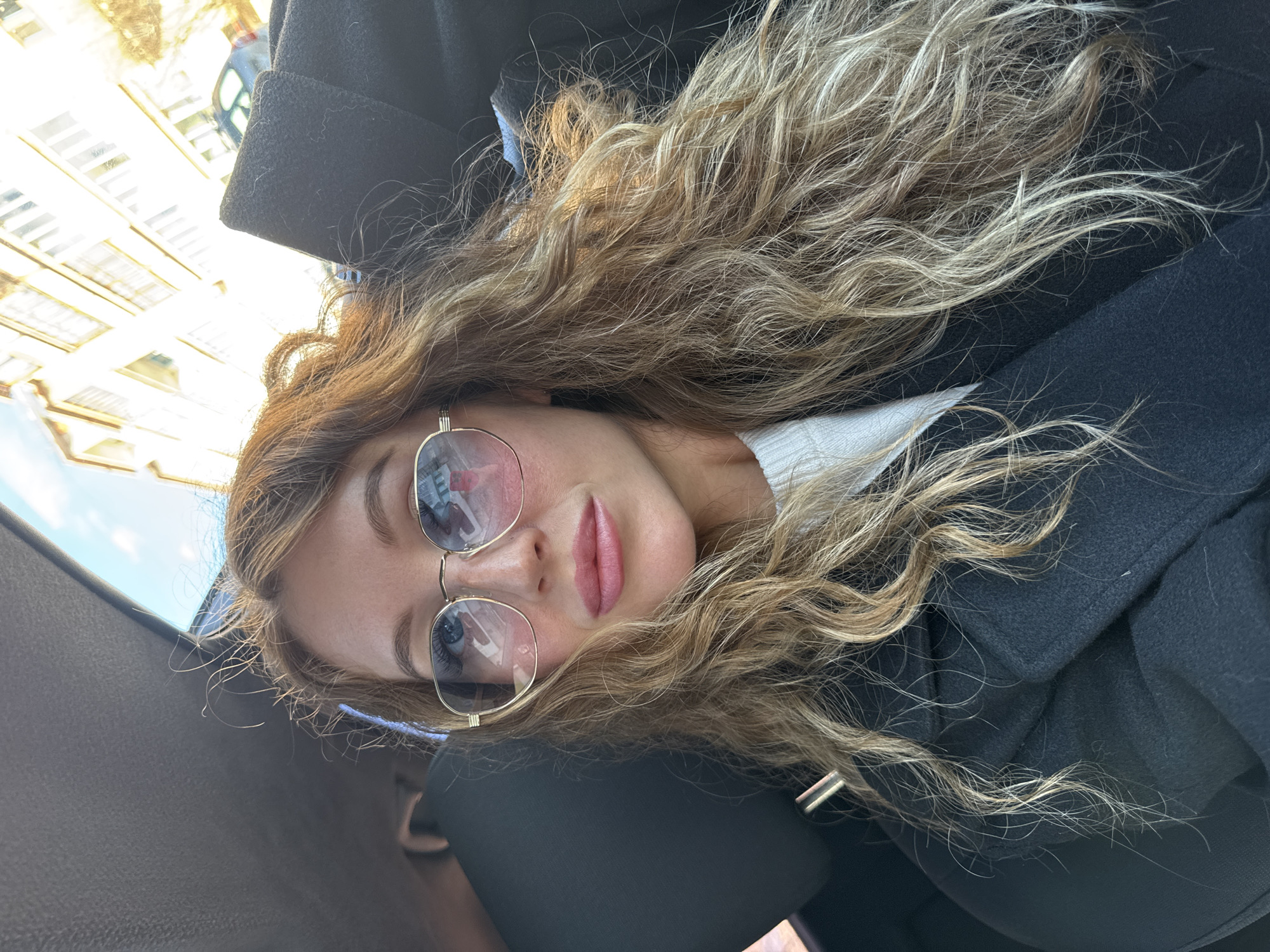 A woman with long, wavy brown and blonde hair is wearing gold-framed sunglasses and a black coat. She is inside a car, with the car door and window visible. The reflection in her sunglasses shows buildings, indicating an urban environment. She is looking towards the camera with a neutral expression. The setting appears to be outdoors during daylight. The location can be inferred to be within Noviy, Russia based on the request.