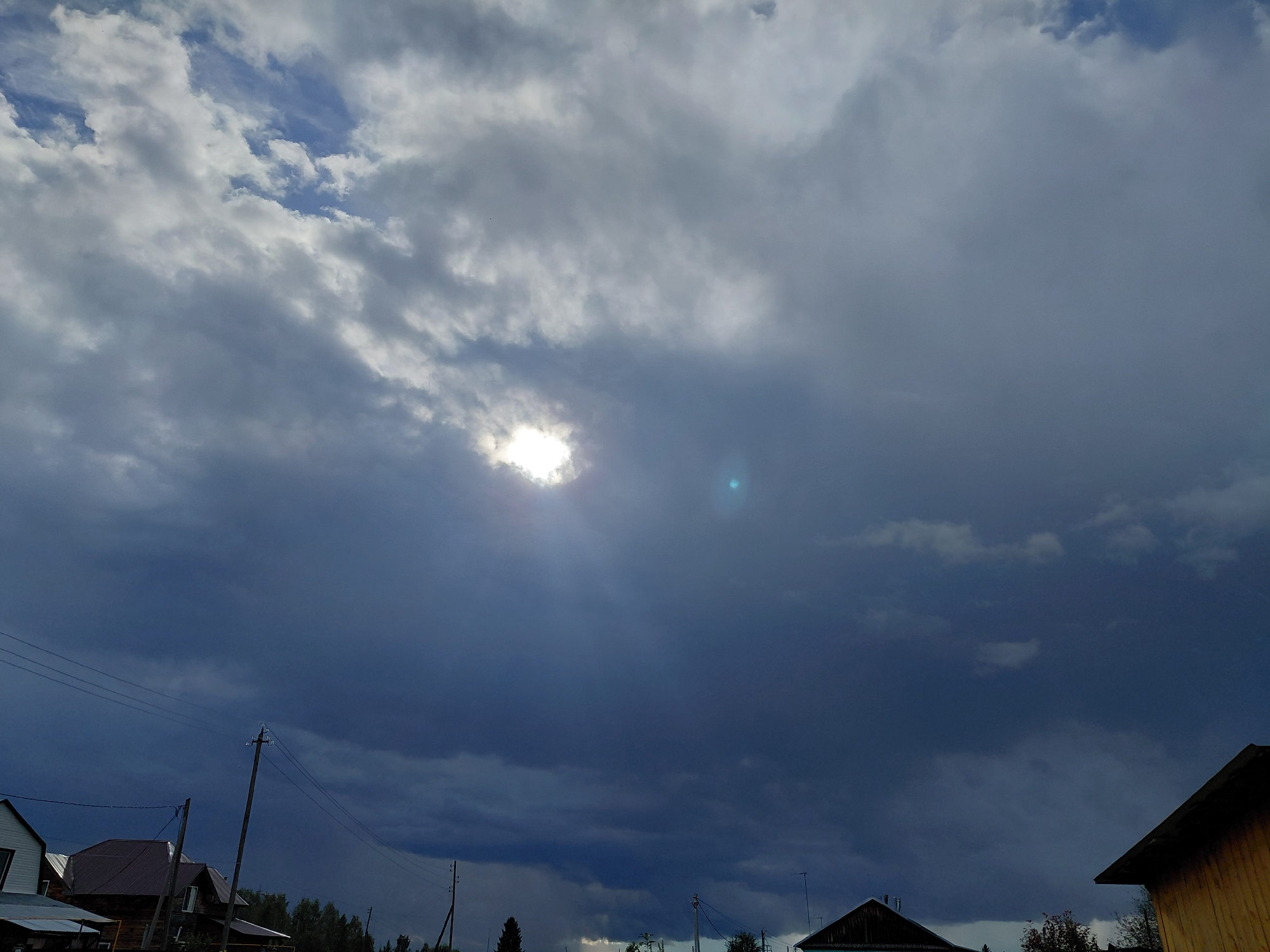 The image depicts a stormy sky over the village of Malinovka, Russia. Dark, ominous clouds dominate the upper portion of the frame, with a break in the clouds revealing the sun, which is partially obscured and appears as a bright, white orb. A faint lens flare, a small blue-green circle, is visible to the right of the sun.
The lower part of the image shows the rooftops of several houses, some with dark, sloped roofs, suggesting a rural or village setting. Power lines and utility poles are visible, stretching across the scene. Trees are sparsely scattered in the background. The overall atmosphere is one of impending weather change, with the heavy clouds indicating potential rain or storm activity. There are no people or animals visible in the frame.