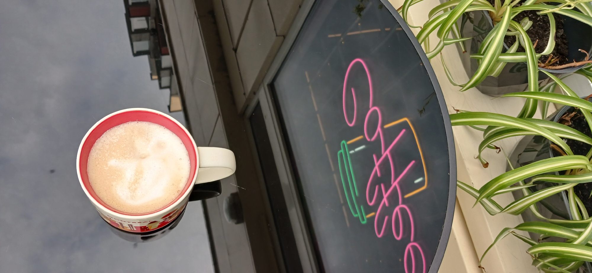 The image captures a close-up view of a glass-topped table with a neon sign reflected on its surface. The sign, in pink and yellow neon, depicts a coffee cup with steam rising. Below the table, a white mug filled with a frothy, light brown beverage, likely coffee with milk, is visible. The mug has a red rim and a colorful wrapper, partially obscured. In the upper portion of the image, lush green spider plants in dark pots are seen. The scene appears to be set outdoors, possibly on a patio or balcony, given the reflections and overhead structures visible in the mirrored surface of the table. The overall ambiance suggests a relaxed, perhaps cafe-like setting. There are no people present in the image, and no specific details point to the city of Khmelnytskyi, Ukraine.