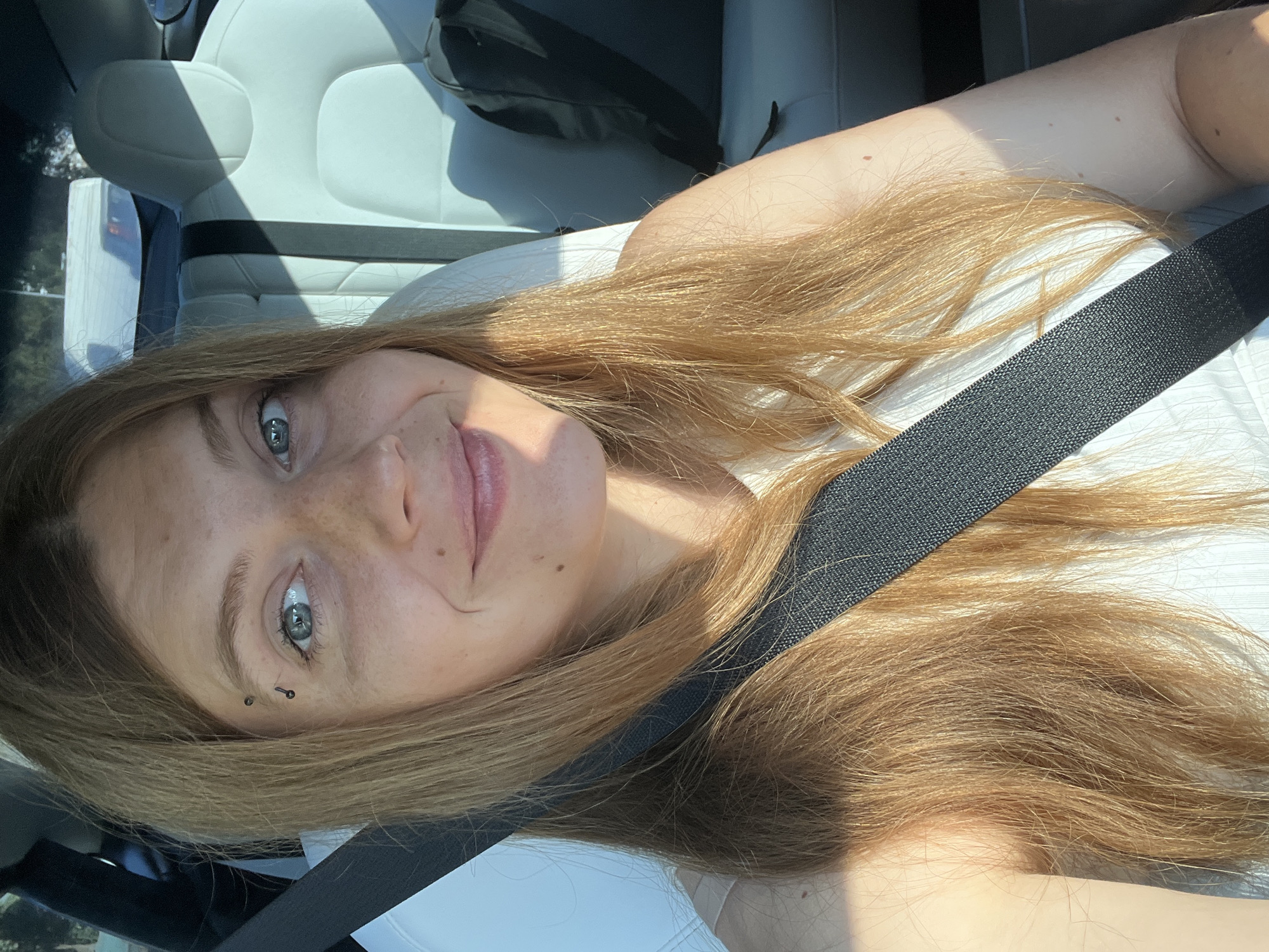 In an indoor setting, possibly a car, a young woman with long, blonde-red hair and light blue eyes is captured in a close-up selfie. She is wearing a white, ribbed top and a black seatbelt across her chest. The seatbelt is prominent in the foreground, with a section of a light-colored car seat visible behind her. She has a subtle smile and her makeup appears natural. There is a piercing above her left eyebrow, consisting of two small, dark studs. Sunlight casts shadows across her face and the interior of the car, suggesting it's daytime. The background is slightly blurred, showing the car's interior and a hint of trees and a white structure outside the window, indicating the car is likely parked or in motion. The image is shot from a medium-low angle, focusing on the woman's face and upper body. The overall impression is casual and relaxed.