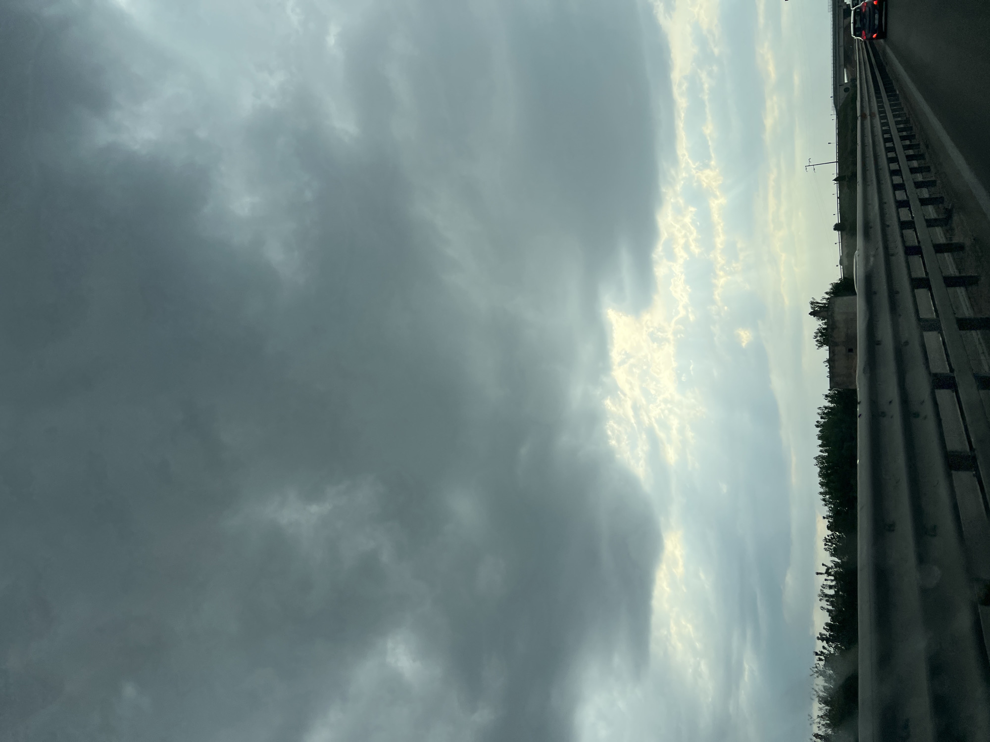 The image captures a view from a moving vehicle on a highway, likely near Trani, Italy, given the context. A dark, overcast sky with heavy, textured clouds dominates the upper portion of the frame. Sunlight pierces through breaks in the clouds in the lower sky, casting subtle rays downwards. Below the sky, a line of dense, dark green trees runs horizontally across the mid-ground, with some indistinct structures or buildings visible behind them. In the foreground, a metal guardrail runs along the side of the highway, indicating the perspective is from within a car or other vehicle traveling on the road. The highway itself is visible, with a portion of a black car in the right rear of the frame, its brake lights illuminated. The overall atmosphere is moody and somewhat dramatic, likely due to the approaching or passing storm.