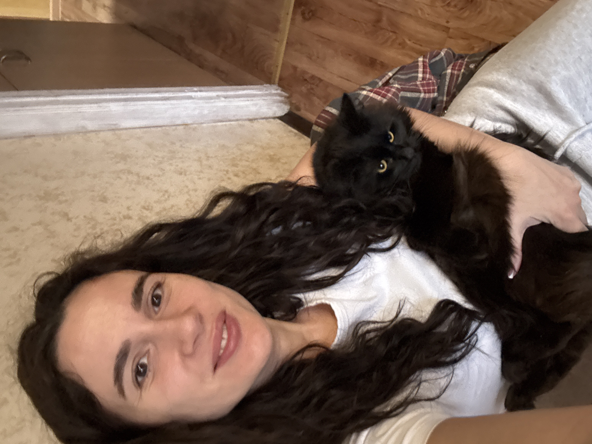 The image captures a close-up, low-angle shot of a woman and a black cat lying together in an indoor setting. The woman, with long, dark, wavy hair spread around her head, is wearing a white t-shirt. She is smiling gently at the camera, conveying a relaxed and affectionate mood. Her left arm is seen gently holding the black cat, which is nestled on her chest or stomach area. Partial view of grey sweatpants is visible on her lower body.
The cat is fluffy with entirely black fur and striking bright yellow eyes, which are directed towards the camera. Part of a red and brown plaid fabric, possibly a blanket or a shirt, is visible behind the cat and the woman's upper body.
The background shows wooden paneling on the walls in the upper sections of the image, and a light-colored, textured floor or carpet beneath the woman. A horizontal, light grey or white strip, possibly a baseboard or part of a piece of furniture, runs across the mid-left background. The lighting is soft and even, suggesting an indoor environment during the day or under artificial light. There is no visible text in the image.