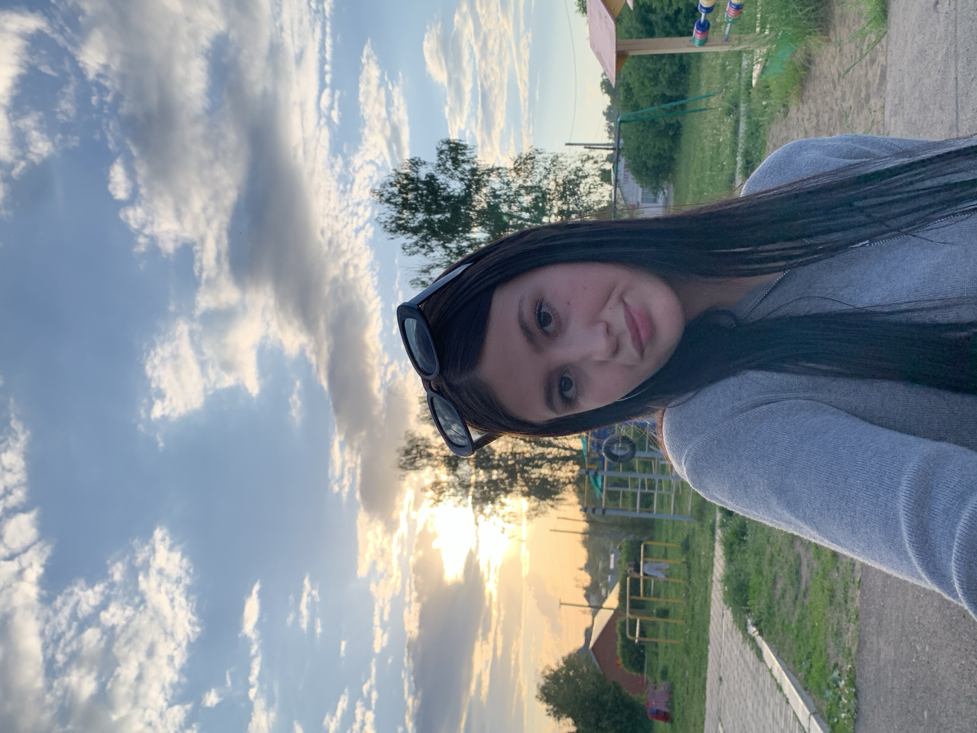 A young woman with long dark hair and sunglasses perched on her head is taking a selfie outdoors. She is wearing a gray sweatshirt. Behind her is a cloudy sky with the sun visible through the clouds. In the background, there is a playground and some greenery. The image appears to be taken in a park in Фоки, Russia.