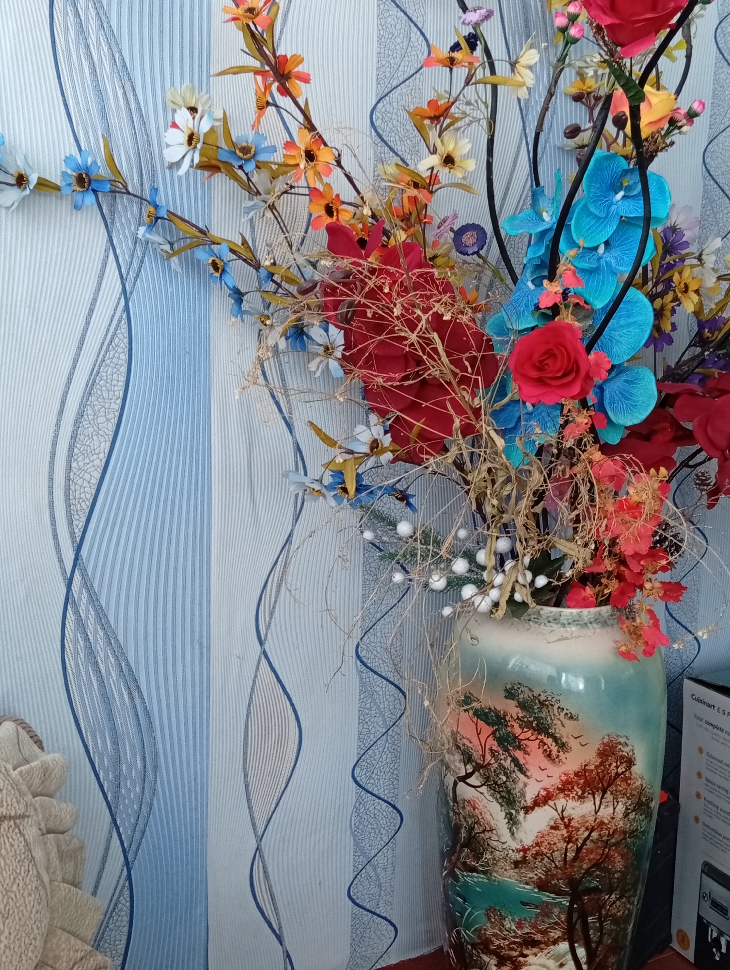 A tall, decorative ceramic vase, painted with a landscape scene of trees and a body of water, stands on a surface in front of a blue and white striped wallpaper with wavy line accents. The vase is filled with a large, colorful arrangement of artificial flowers and dried branches. The flowers include red roses, bright blue orchids, and various smaller blooms in shades of orange, yellow, and white. Some of the dried elements appear to be twigs and small white bead-like decorations. To the right of the vase, a portion of a white box with text and an image of a coffee machine is visible. The overall scene suggests an indoor setting, likely a home or a decorative display area. The location is unknown.