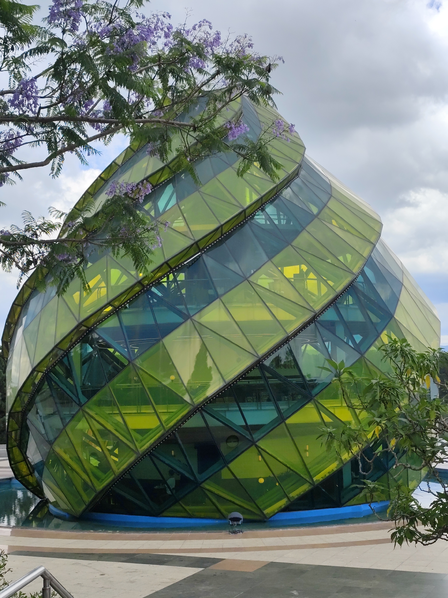 The image shows a unique, geometrically-designed building in Da Lat, Vietnam, known as the Cafe Bien Tau. The structure is primarily composed of green and yellow glass panels arranged in a spiraling, ovoid shape. A small body of water surrounds the base of the building, reflecting its image. The foreground and portions of the background are obstructed by the branches of flowering trees, displaying lavender blossoms. The sky is overcast with grey clouds. A metal railing is partially visible in the lower-left corner, suggesting a public viewing area. A small utility box is visible in front of the structure, near the water.