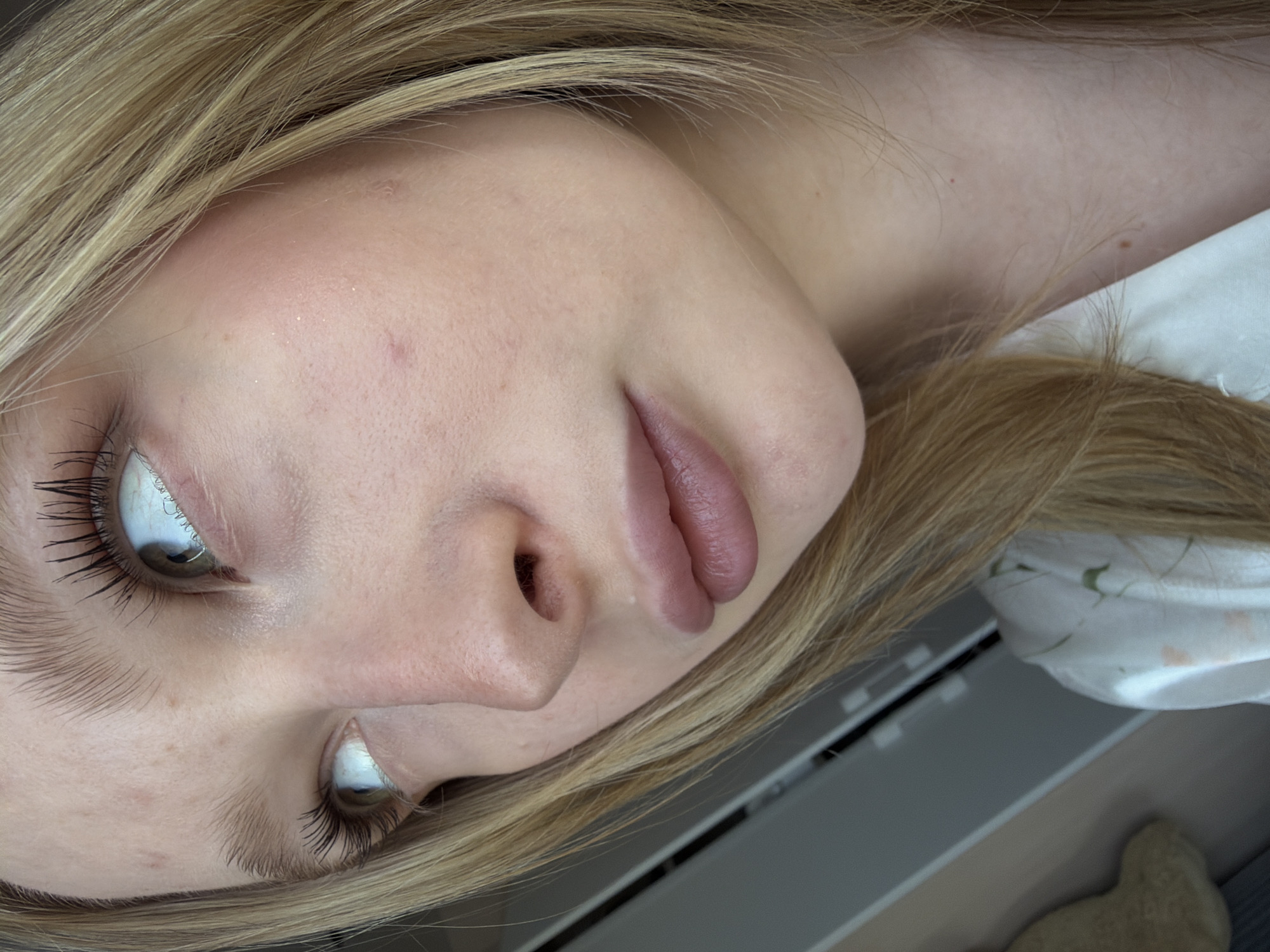 A close-up photograph shows a light-skinned person with blonde hair, tilted at an angle. The person's face is the primary focus, featuring eyes with mascara, a nose, and lips. The background includes a white fabric with a floral pattern and a grey surface that could be a desk or other furniture. The location of the photo in the city of Rossiyskiy, Russia is not discernible from the image.