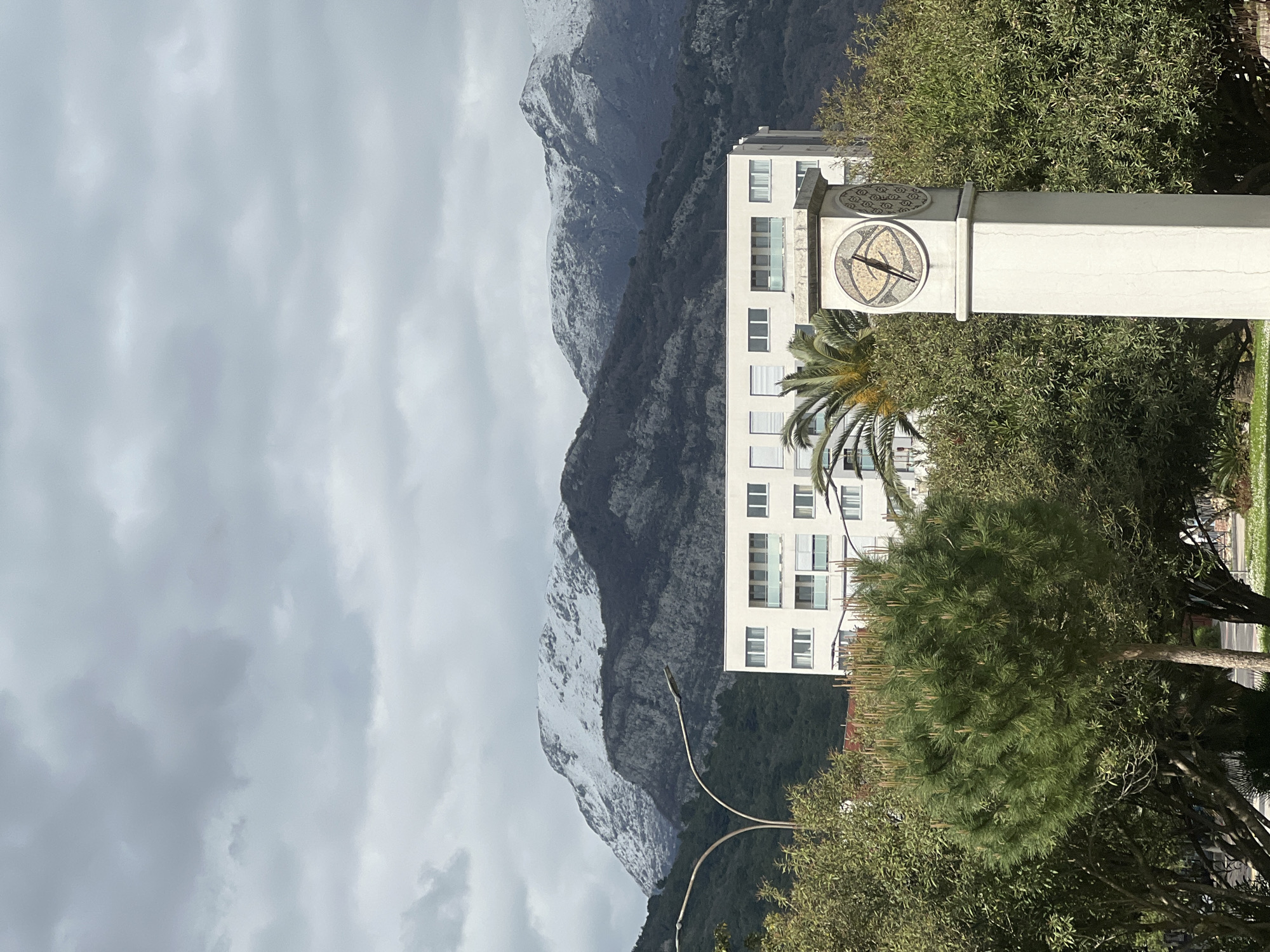 The image shows a scene in Budva, Montenegro, featuring a prominent clock tower situated amidst green trees and foliage. In the background looms a large mountain range with visible patches of snow. To the left of the clock tower stands a modern, multi-story white building with multiple windows and balconies. The sky is overcast with grey clouds, providing soft, diffused lighting across the scene. A palm tree is partially visible beside the clock tower. In the foreground, various trees and greenery fill the lower portion of the image. A street lamp is partially visible. The focus is sharp, capturing the details of the clock face, the building's architecture, and the rugged terrain of the mountains.