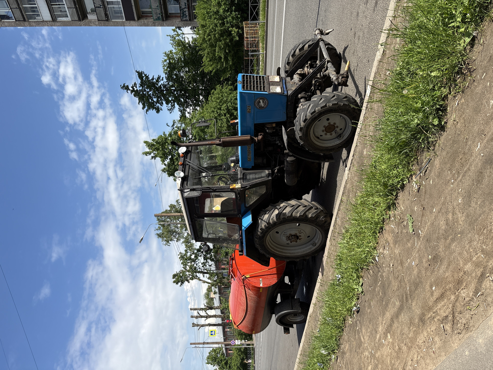 The image, viewed upright, depicts a blue Belarus MTZ-82.1 tractor parked on the side of an asphalt road under a bright, sunny sky. The tractor is equipped with a large, orange, cylindrical tank attachment mounted at its rear, suggesting its use as a municipal watering or spraying vehicle.
The vehicle is positioned next to a concrete curb, bordering a strip of green grass and exposed dirt. In the background, a row of green trees lines the street, partially obscuring a multi-story building with multiple windows on the left. The sky is a clear blue with scattered white, wispy clouds. Strong sunlight casts distinct shadows, indicating midday or early afternoon.
Further down the street, utility poles with overhead wires and streetlights are visible, along with a yellow triangular road sign, likely a warning or information sign for drivers. The overall setting is an urban environment.
Visible text on the side of the tractor includes "82.1" and the Cyrillic word "БЕЛАРУС" (BELARUS), identifying the model and manufacturer. No people are visible in the scene. The architecture and equipment are consistent with the provided location context of Saint Petersburg, Russia.