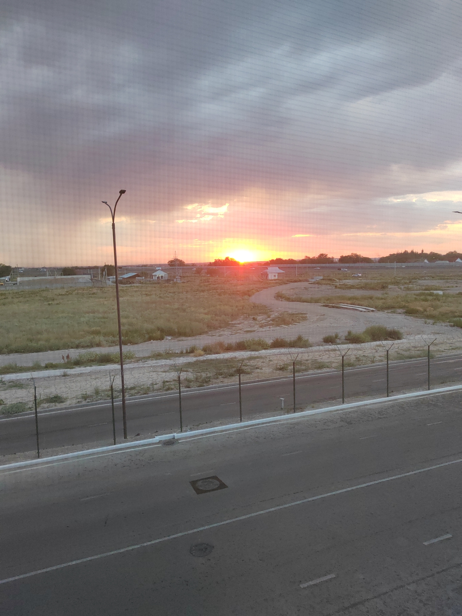 The image captures a sunset scene in Isaevo, Kazakhstan. The sky is filled with dramatic clouds, tinged with the warm hues of the setting sun, creating a soft glow. In the foreground, a paved road stretches across the frame, marked with white lines. A fence with barbed wire runs along the edge of the road, separating it from a field of dry grass and scattered construction materials. A single street lamp stands tall near the center of the image, its silhouette stark against the colorful sky. In the distance, a few buildings and cars are visible, suggesting a sparsely populated area. The overall impression is one of a quiet, rural landscape bathed in the golden light of dusk.