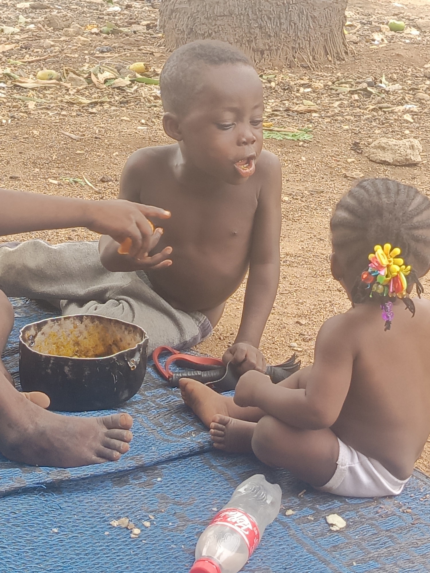The image shows three children seated on a blue mat outdoors. One child, shirtless, is eating from a bowl held by another child whose hand is visible. A second child, also shirtless and wearing a white diaper, sits facing away from the viewer, with colorful hair accessories. A bottle of Coca-Cola lies on the mat. The background consists of sandy soil with scattered leaves, suggesting an unpaved area. The location is in Akwanga, Nigeria.