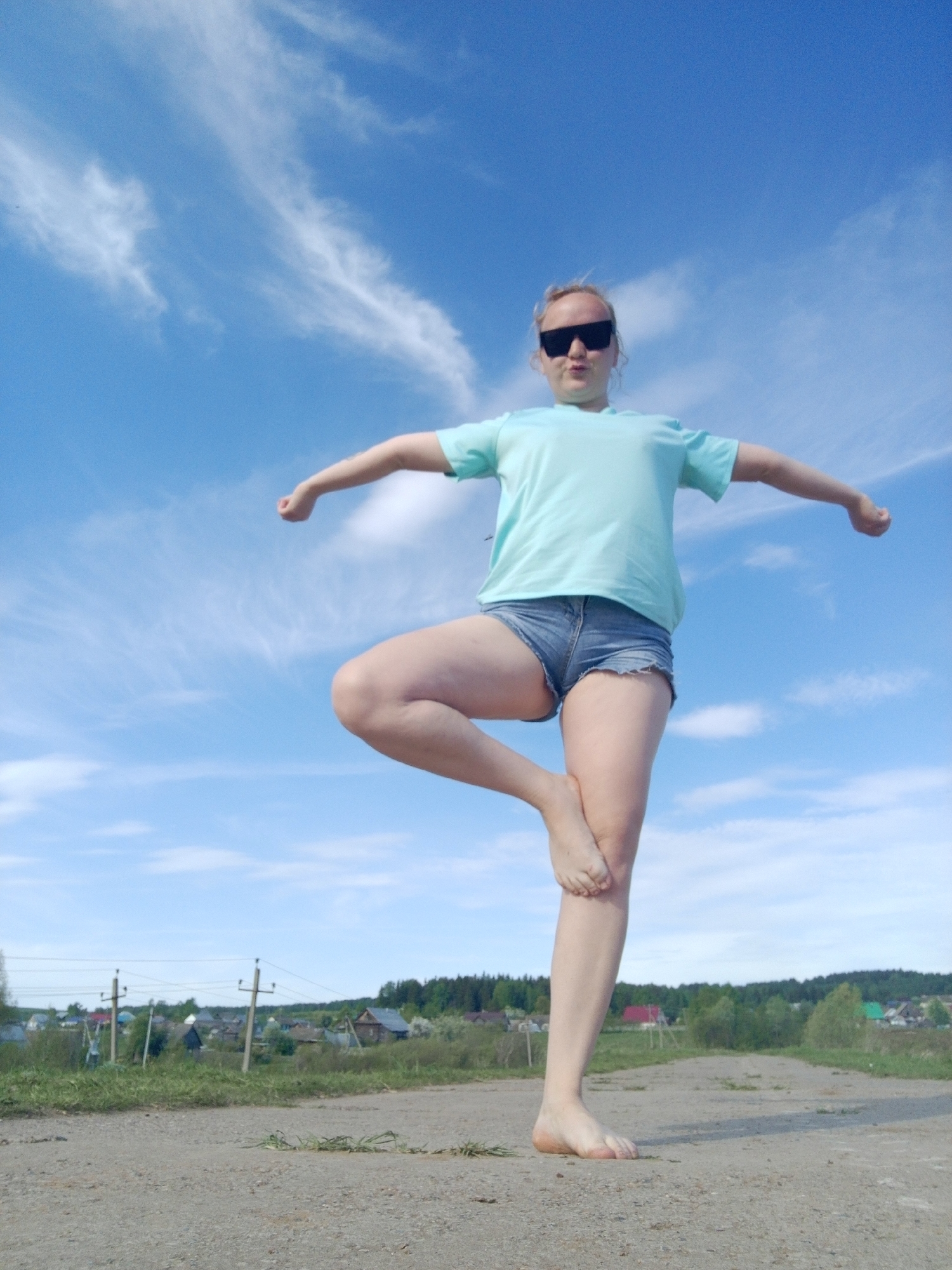 A woman is standing on a dirt road in Уральское, Russia, balancing on one leg. She is wearing a light blue t-shirt, jean shorts, and dark sunglasses. Her arms are outstretched to the sides. In the background is a blue sky with scattered clouds, a grassy area, and houses.