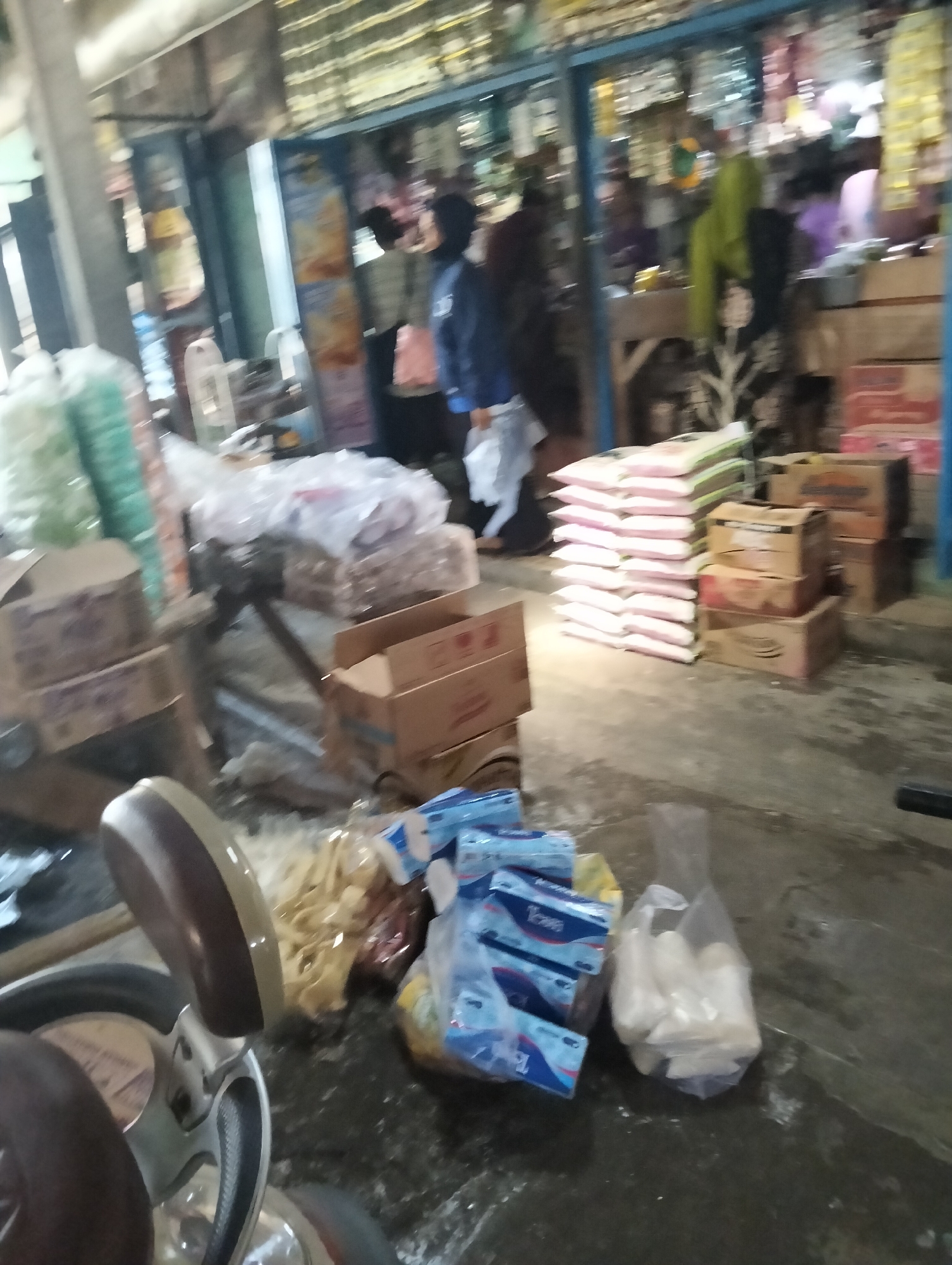The image depicts a busy market scene in Muncar, Indonesia. Stacks of goods, including what appear to be bags of rice or flour, are arranged on the ground and on shelves. Cardboard boxes are also present, some open and displaying their contents. Several people are visible, but their faces are obscured by distance or the quality of the image, making it difficult to identify them. One person is wearing a blue jacket and carrying a white bag. Another person is seen in the background wearing a green headscarf. Bags of packaged snacks and toiletries are also scattered around the market floor. The overall environment suggests a typical outdoor market or a small shop with goods displayed for sale.