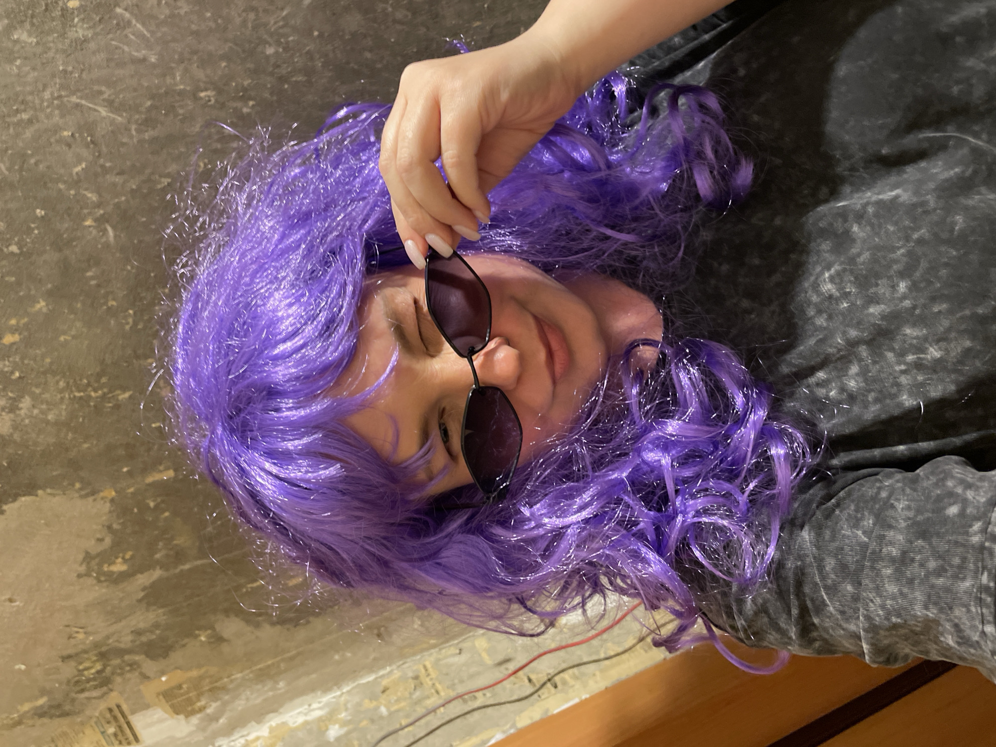 A person with purple hair is lying down, holding sunglasses. The person is wearing a grey shirt. The floor is mottled grey and brown. Two thin wires (red and black) are visible in the lower left. There is no information in the image to indicate the location is in Izhevsk, Russia.