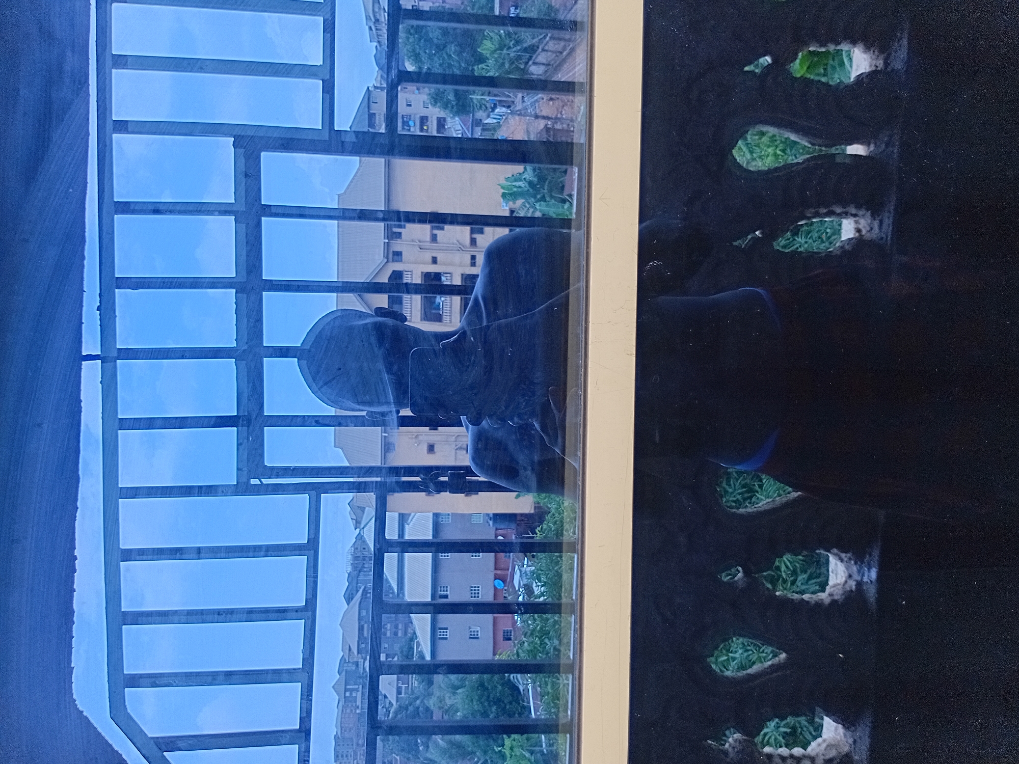 The image captures a reflection in a window, looking out at a cityscape in Nkpor, Nigeria. The foreground of the reflection shows the silhouette of a person holding a phone to take a picture, with their head and shoulders visible. Below this reflection, a decorative, dark railing with an ornate pattern is visible, framing a view of lush green foliage.
The scene beyond the window displays a densely populated urban environment. Multiple multi-story buildings, some with light-colored facades and others with darker exteriors, are clustered on a hillside. The architecture suggests a mix of residential and possibly commercial structures. Some buildings have visible windows and balconies. The sky is overcast with muted tones, indicative of a cloudy day. In the middle ground, there are trees and some vegetation interspersed with the buildings, suggesting a somewhat developed but still green urban area. A water tank is visible on top of one of the buildings. The overall impression is of a typical Nigerian city during daylight hours, with a sense of depth created by the layers of buildings and the surrounding landscape.