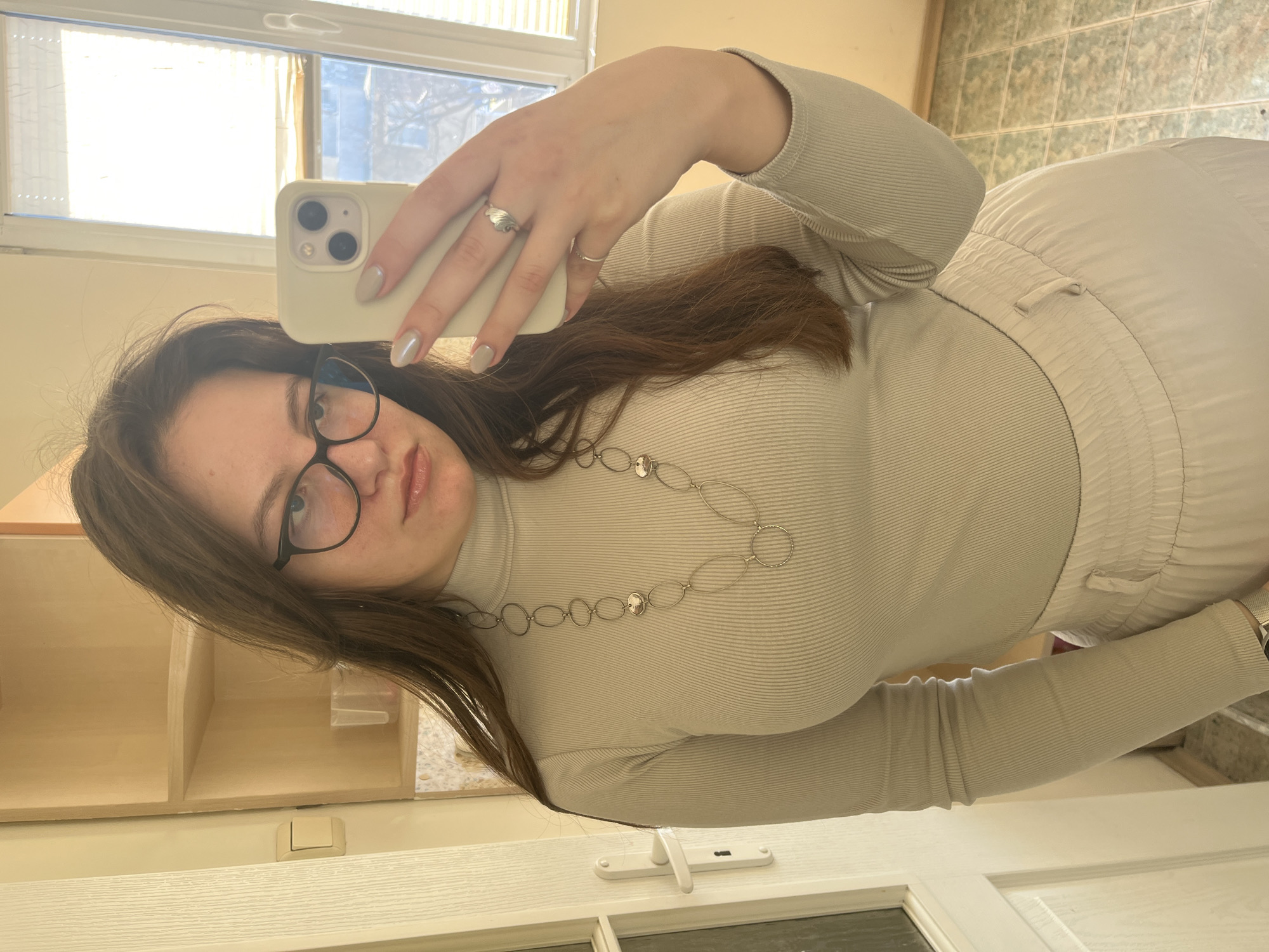 A female subject is depicted in a mirror selfie taken indoors in Varna, Bulgaria. She has long brown hair, wears black-rimmed glasses, a light-beige ribbed turtleneck top, and light-colored pants. A silver-toned necklace with oval and circular links is worn, and two silver rings adorn her right hand, which is holding a light-colored smartphone to capture the image. Her nails are manicured with light-colored polish. The background includes a white door with a handle and keyhole on the left, a light-toned shelving unit with a clear plastic cup visible on a shelf, and a window with horizontal blinds on the right. Through the window, faint outlines of urban buildings are visible. The floor is covered with light green and off-white checkered tiles.