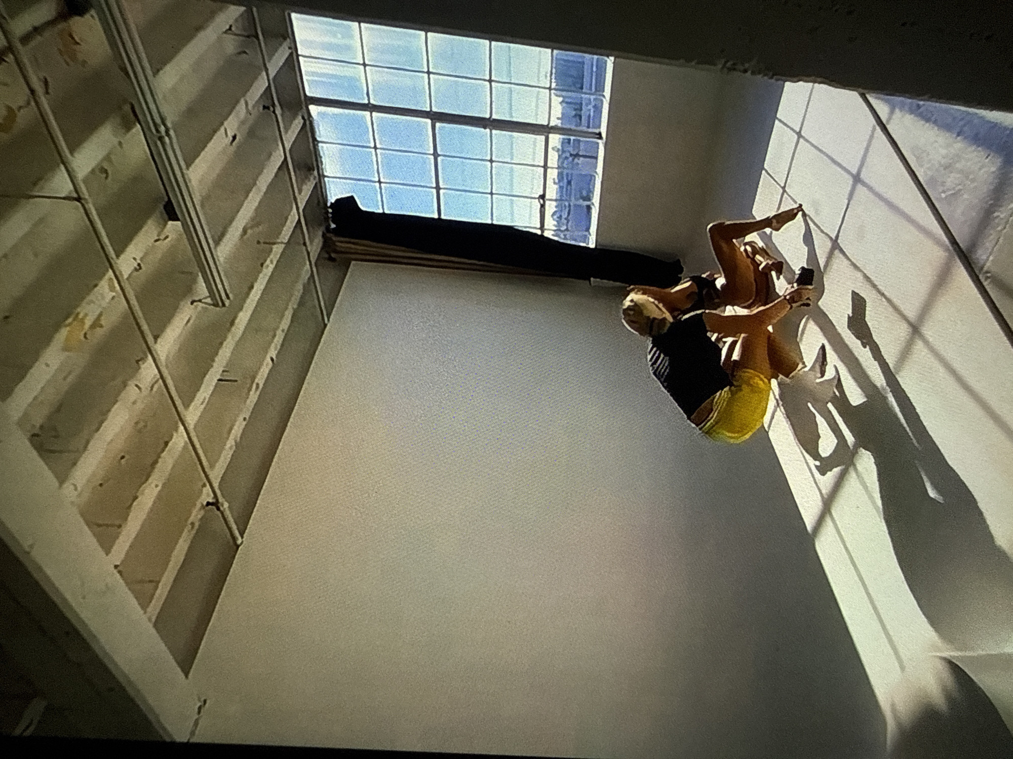 The image displays an indoor scene, likely a photography studio or industrial loft, located in Varna, Bulgaria. The room features plain white walls on the left and rear, forming a seamless background that transitions into a matching white floor. The ceiling consists of exposed concrete beams with several suspended fluorescent tube light fixtures. A large, multi-pane industrial-style window occupies the right wall, admitting strong natural light that casts distinct grid shadows from the window frame onto the floor and lower wall, along with sharp shadows of the subjects. A dark curtain hangs along the left side of the window.
Through the window, a distant urban landscape is visible under a clear blue sky, comprising various buildings and structures, suggesting an expansive city view, possibly near a body of water.
Two individuals are present in the lower right-center of the frame. The individual on the left has blond hair, is wearing a black short-sleeved top, bright yellow shorts, and white footwear. She is crouched down, facing the second individual, and appears to be holding a dark-colored smartphone or small camera in her right hand, casting a clear shadow on the floor. The second individual, positioned to the right, has lighter hair and is wearing minimal dark-colored attire, resembling a swimsuit or lingerie. She is posing on the floor, leaning back on her left arm with her legs bent. Their proximity and actions indicate a photographic session.