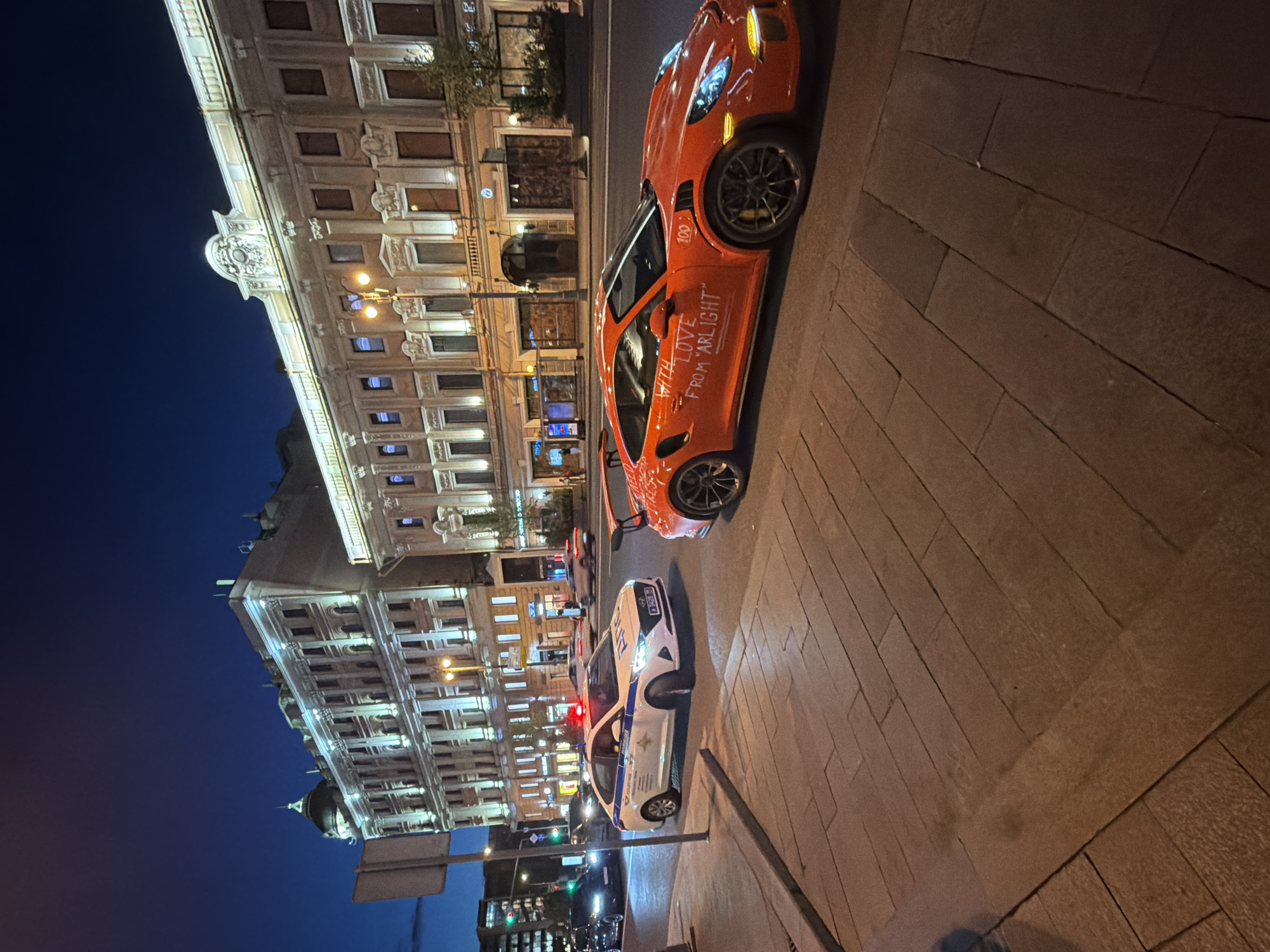 The image captures a nighttime street scene in Moscow, Russia, characterized by a blend of illuminated architecture and modern vehicles. In the foreground, an eye-catching orange sports car, identifiable as a Porsche (likely a 911 GT3 RS), is parked or stopped on a paved surface. Its side features the distinct white text "WITH LOVE FROM ARLIGHT" and "#722_OVL", along with a "100" decal, suggesting a personalized vehicle or participation in an event.
Adjacent to the sports car is a white Russian police vehicle, clearly marked with "ДПС" (DPS, State Road Safety Inspectorate) in blue lettering, and exhibiting blue flashing lights. The presence of both vehicles side-by-side suggests a typical urban environment, possibly a routine stop or just passing traffic.
The background is dominated by impressive, multi-story buildings with classical, ornate architectural details, brightly lit, which stand out against the deep blue to black night sky. Streetlights cast a warm glow on the pavement and surrounding area. Further down the street, other vehicles and traffic lights (showing both green and red signals) are visible. The ground is covered with dark, rectangular paving blocks. The overall atmosphere is that of a bustling city evening, with no specific human activity or emotion prominently displayed.