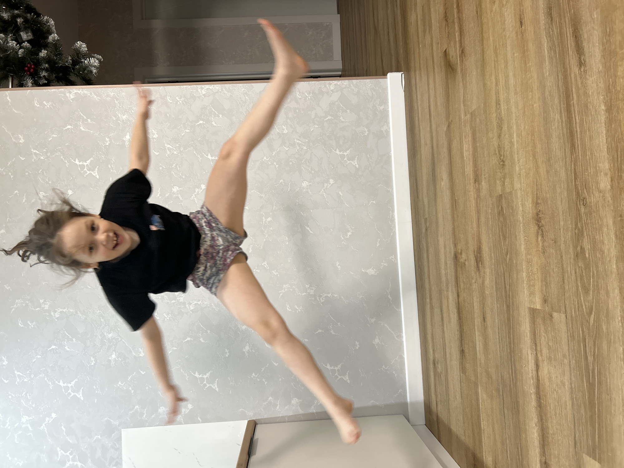 A young child is photographed mid-air. The child is wearing a black top and patterned shorts. The child's right leg is extended upward and slightly to the right, while the left leg is extended downward. The child's hair is loose and moving.
The background features a light gray wall with a marble-like pattern. The floor is a light brown wood-grain pattern. A Christmas tree is visible in the top left corner.
The photograph was taken inside a house in Chaikovsky, Russia.
