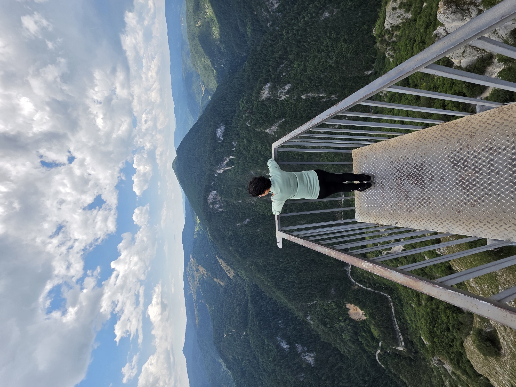 A person stands on a metal viewing platform, leaning on the railing, and looking out over a vast, verdant mountain landscape. The person, seen from behind, has dark, curly hair and is wearing a light green long-sleeved shirt and dark pants. The viewing platform is constructed of diamond plate metal flooring and a metal railing. Below, the landscape consists of steep, tree-covered slopes and deep ravines, with a winding road visible in the distance. The sky above is a mix of blue sky and fluffy white clouds. This viewpoint is located at the Eagle's Eye (Ochite na Orlite) panoramic platform in the Rhodope Mountains, near the village of Yagodina, Bulgaria.