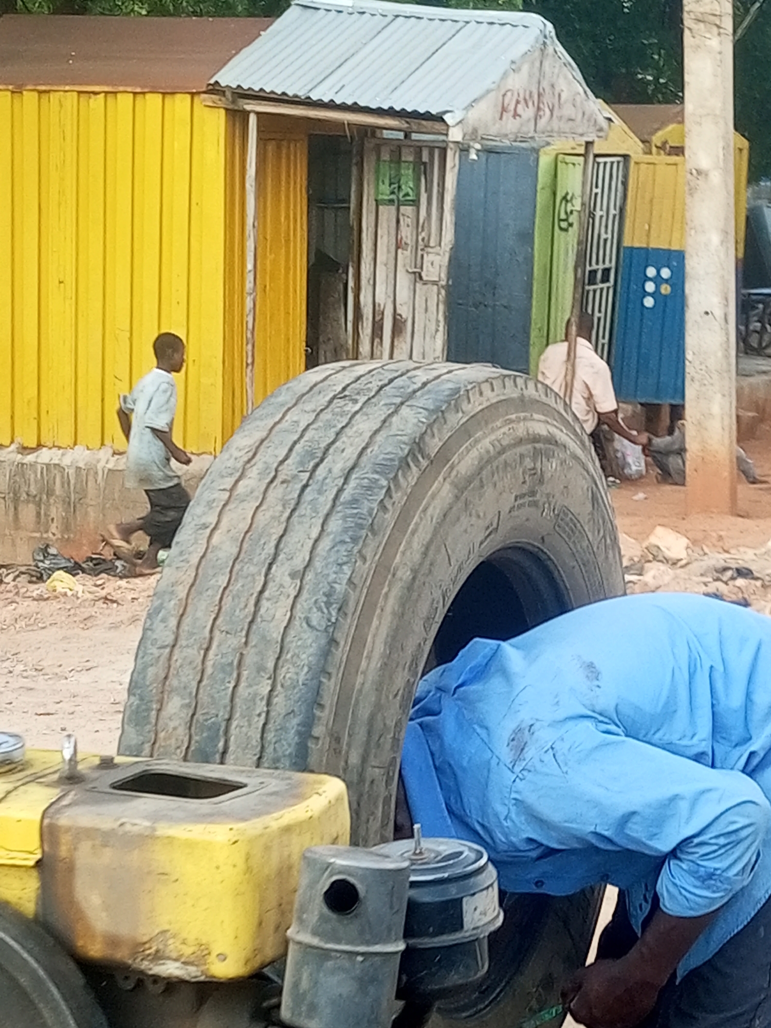 The image depicts an outdoor scene with several elements. In the foreground, a large, worn tire dominates the lower part of the frame. Partially visible behind it is a person wearing a light blue shirt, leaning over to work on something, their back to the camera. A yellow object, possibly part of a machine, is to the left of the tire.
In the mid-ground, a young boy in a light t-shirt and dark pants walks away from the viewer, heading towards a row of colorful structures. Behind him, the structures appear to be makeshift buildings or stalls with corrugated metal walls in yellow, blue, and green. One structure has a sign with red lettering that is partially obscured but seems to read "PEMBYL". Another has a blue door with white circles arranged in a pattern.
In the background, more of these structures are visible, along with some greenery suggesting an outdoor environment. The ground is sandy and appears somewhat unkempt, with some debris scattered around. The lighting suggests it could be daytime, possibly late morning or afternoon, with no clear indications of specific weather conditions. The overall scene suggests a busy, possibly informal commercial area or a workshop.