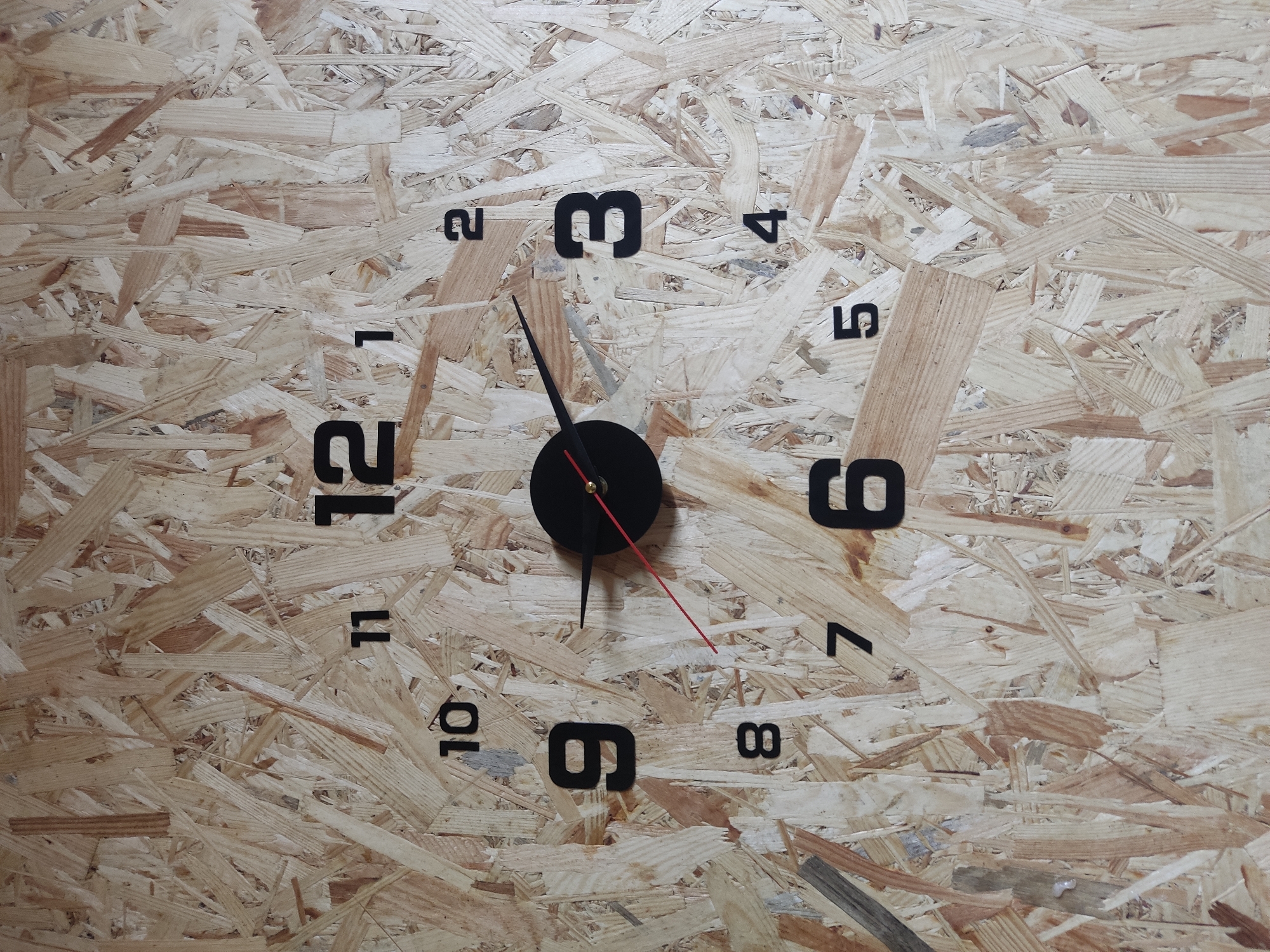 This is a close-up, vertical, eye-level shot of a wall clock. The clock face is made of oriented strand board (OSB), a wood-based panel material. The numbers indicating the hours are black and are affixed to the OSB at their respective positions. The hour hand is black and points slightly past the 9, and the minute hand is also black and points to the 12. A thin red second hand points to approximately the 5. The clock is circular and appears to be a decorative wall-mounted item. There is no indication of a city, country, or specific location within Radisne, Ukraine. The image focuses solely on the clock and its immediate background.