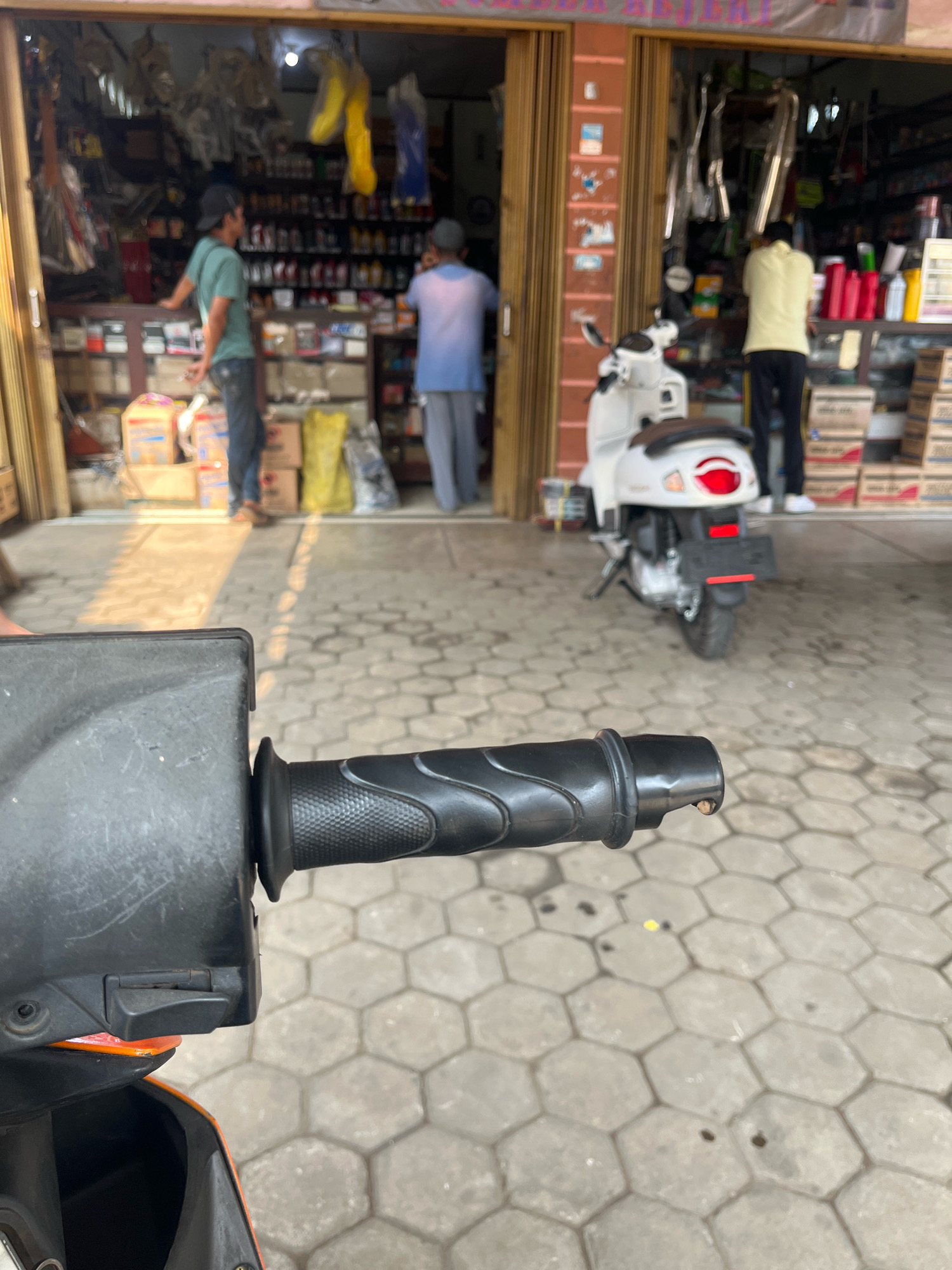 In Kemuning, Indonesia, a close-up view of a motorcycle's handlebars dominates the foreground. The handlebars are black and textured, with a grip that shows signs of wear. Behind the handlebars, a white scooter is parked on a hexagonal-patterned paved surface. The scooter has a brown seat and a red taillight. To the left of the scooter, a shop entrance is visible, with shelves stocked with various items, including bottles and boxes. Two men are present in the shop. One man, wearing a blue shirt and gray pants, is standing and looking towards the shelves. Another man, wearing a light yellow shirt and black pants, is also in the shop. The shop appears to be an automotive parts store, with exhaust pipes hanging from the ceiling. Sunlight casts shadows across the pavement in front of the shop.