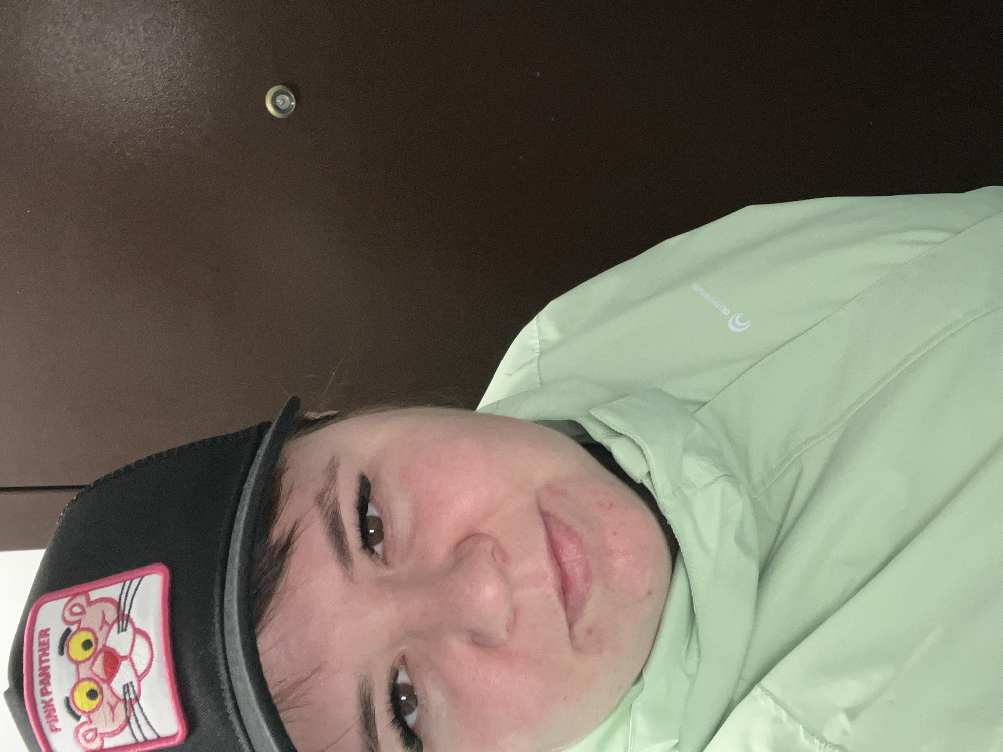 A woman with fair skin and brown hair is shown from the neck up, wearing a light green jacket and a black baseball cap with a Pink Panther patch. She is facing the camera at a slight angle and has a neutral expression. The background is a dark brown, possibly a door or wall, with a peephole visible in the upper left corner. There is no location in the city of Chaikovsky, Russia.