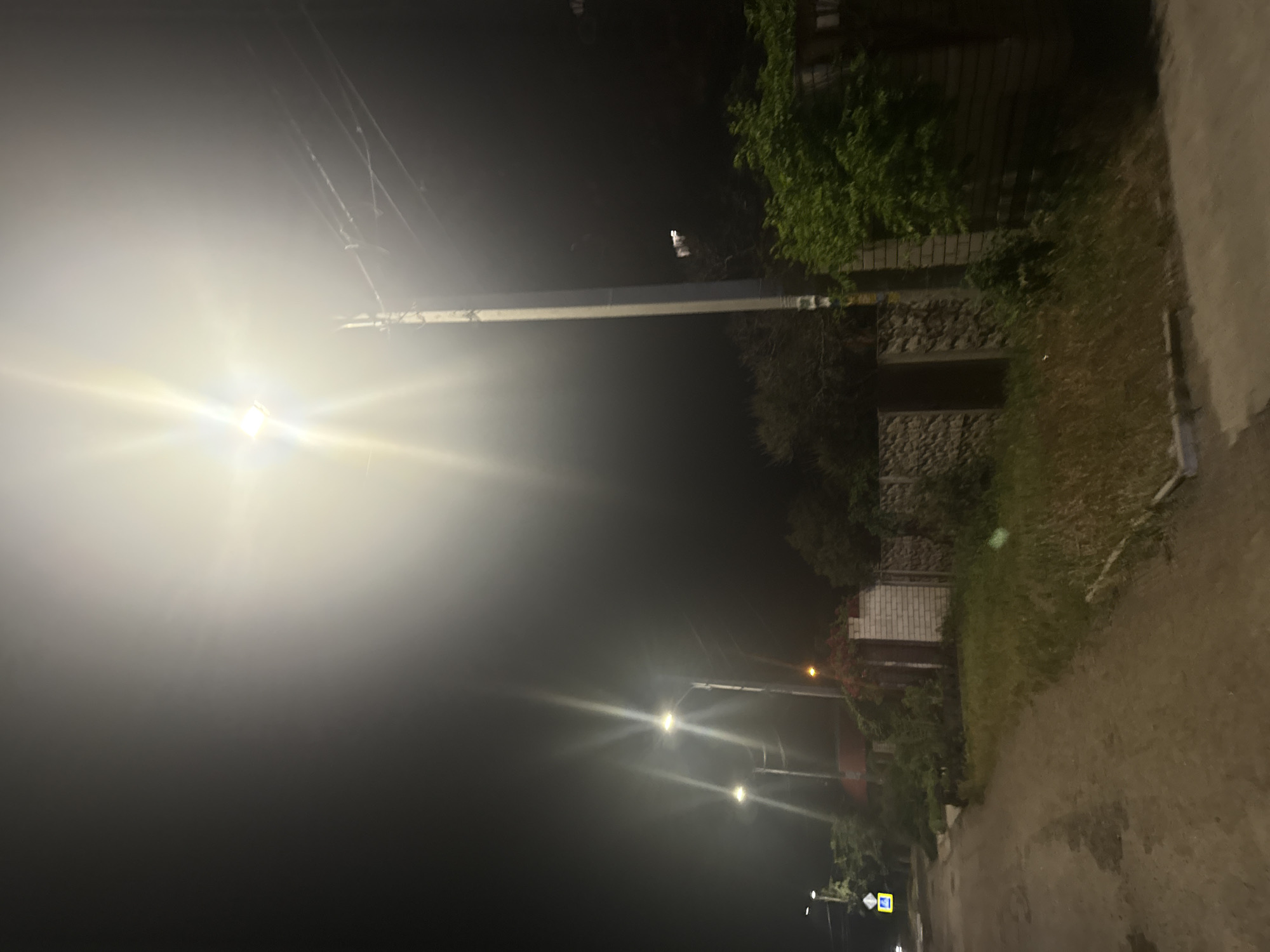 This image depicts a nighttime street scene in Dnipro, Ukraine. The primary light source is a bright, overexposed street lamp positioned in the upper right quadrant, casting a strong glare that obscures details in its immediate vicinity. To the left of this main light, another street lamp is visible, also producing a bright beam. The scene is characterized by a dark, hazy sky, indicative of a poorly lit environment or atmospheric conditions like fog or smoke.
In the foreground, a dirt road or pathway runs from the lower left towards the center of the image, showing some unevenness and patches of darkness. Along the right side of the road, a low stone fence and a brick wall are visible, with dense green foliage growing over and behind them. A wooden fence also borders the property on the right. Several street lamps are interspersed along the street, with one on the far left appearing to be a standard traffic signal or directional marker with a yellow diamond shape and a white arrow.
The overall impression is one of a quiet, possibly rural or suburban street at night, illuminated by artificial lighting. The high contrast between the bright lights and the dark surroundings, along with the lens flare, creates a somewhat atmospheric and potentially disorienting effect. No individuals are present in the frame.