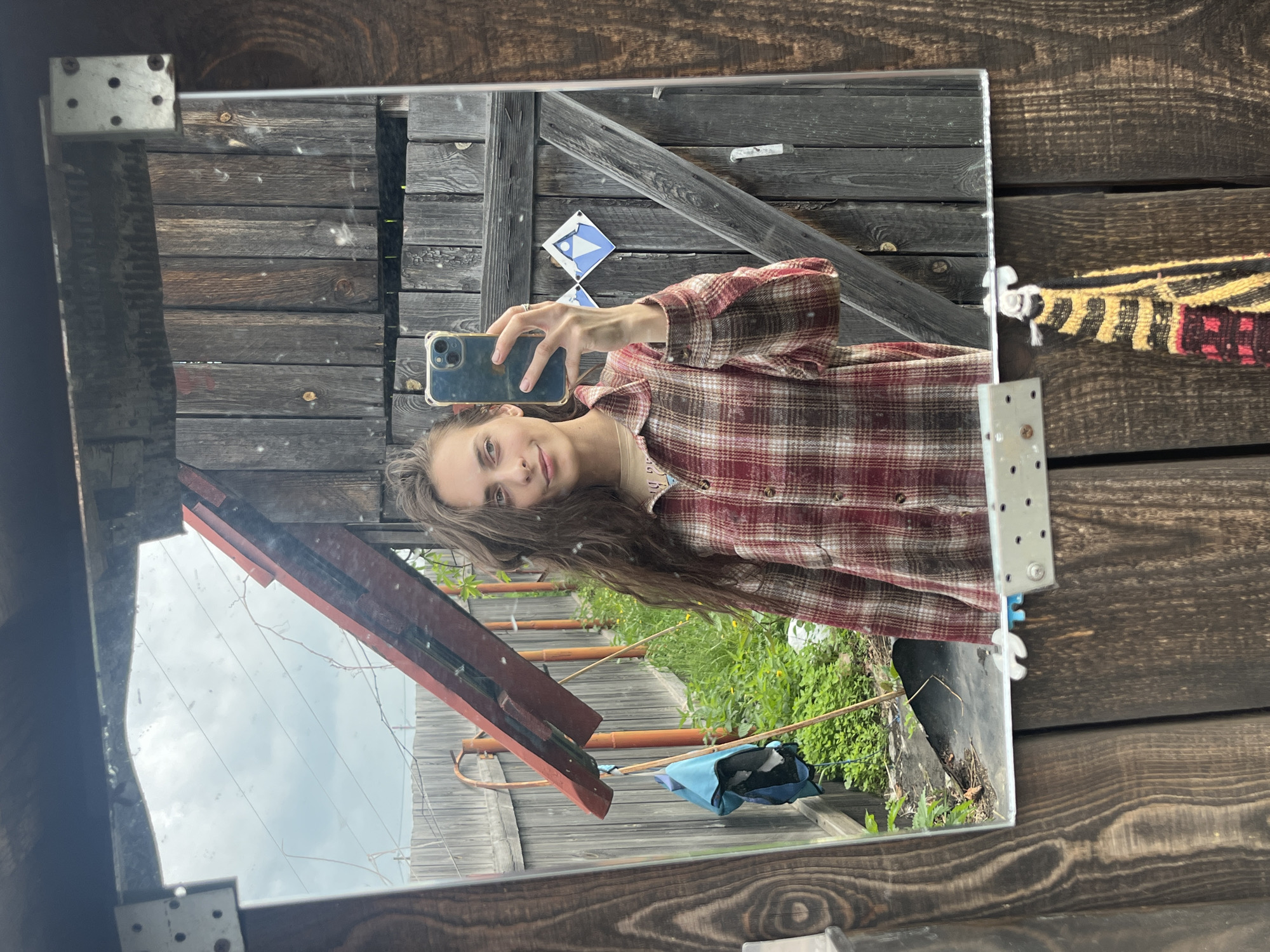 A woman takes a mirror selfie outdoors. She has long, dark, wavy hair and is wearing a red and brown plaid shirt over a tan t-shirt. She holds a smartphone in her right hand to capture the reflection. The background behind her is a rustic wooden fence and some greenery. The mirror itself is framed by weathered wood. The setting appears to be a yard or backyard, likely in Berezovskii, Russia, given the wooden structures and natural surroundings.