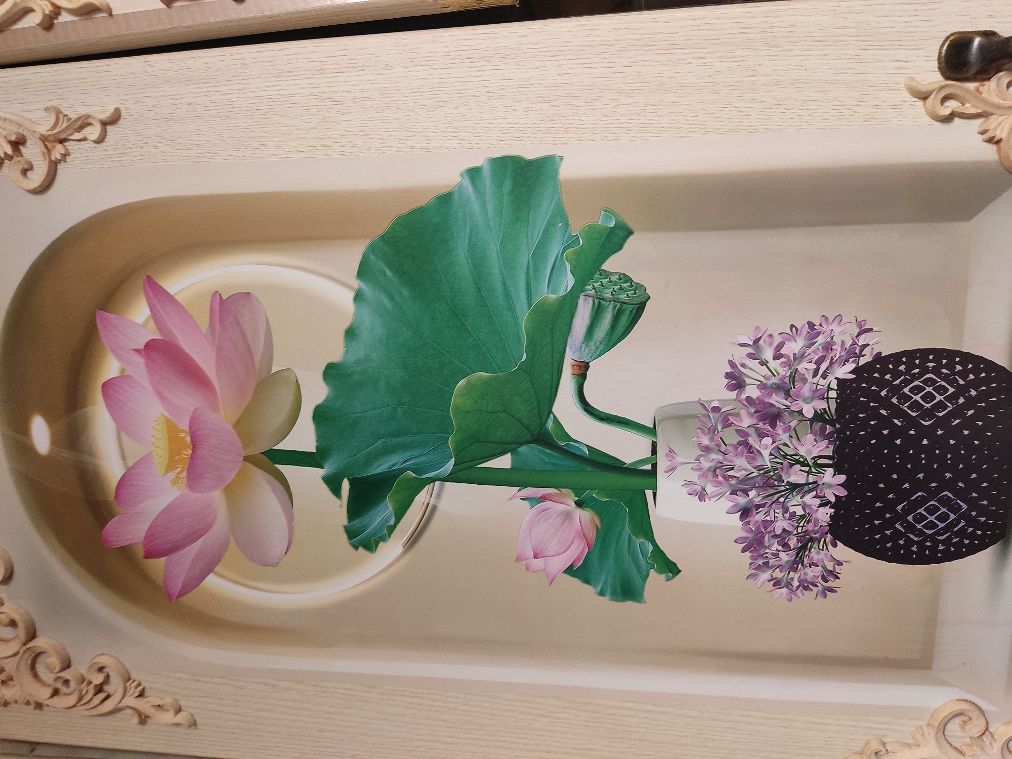 The image shows a decorative panel featuring a still life of lotus flowers and smaller purple flowers in vases, set against a backdrop of an archway. Intricate wooden carvings adorn the top and sides of the panel. The scene is static and appears to be a piece of interior decor. There are no people, actions, or interactions visible. The location is not specified in the image itself.