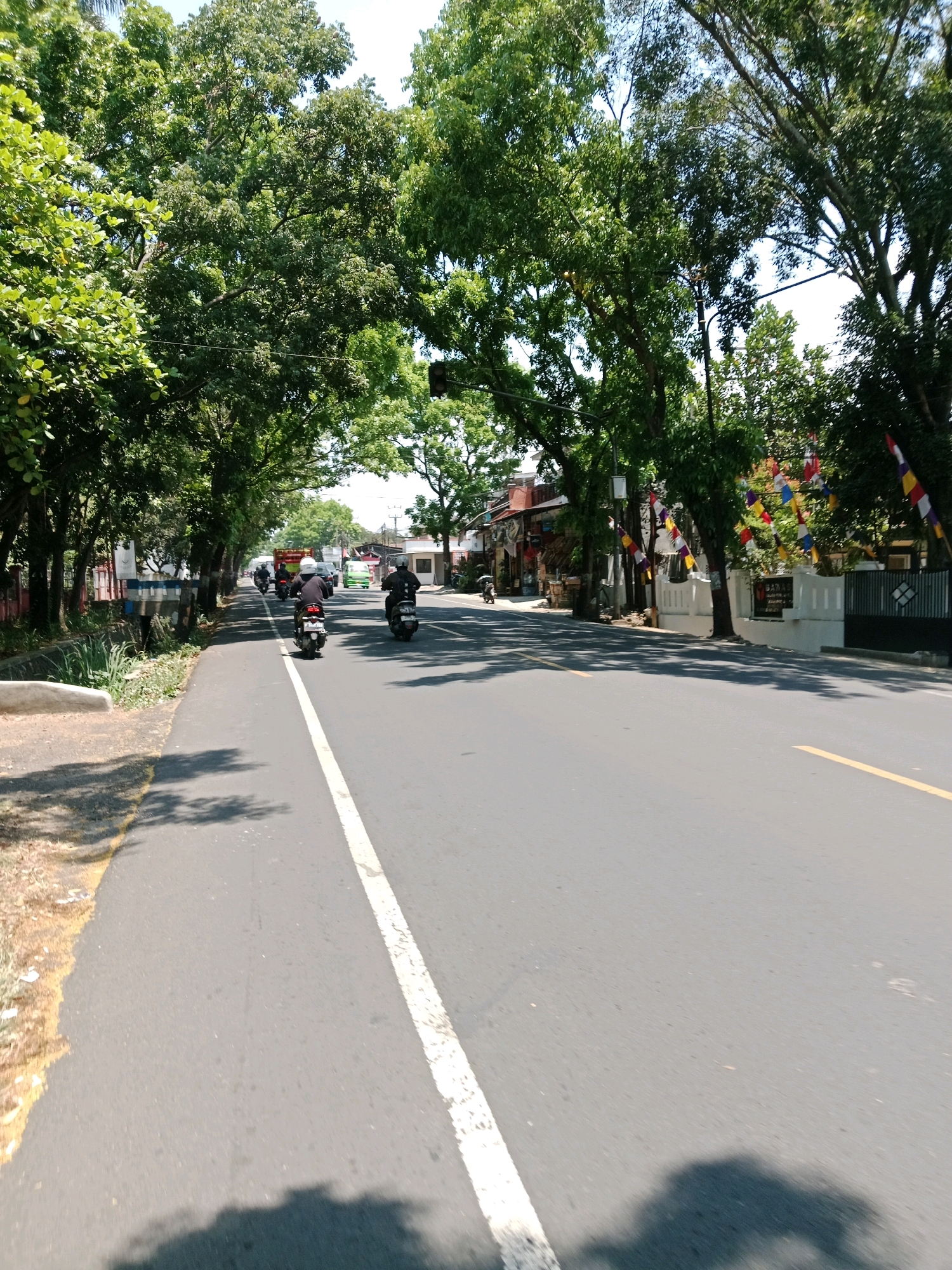 This is a photograph taken on a sunny day, showing a street scene in Cianjur, Indonesia. The perspective is from a low angle, suggesting the photographer is either walking or riding a vehicle. The street is paved with asphalt and has a white line marking the lane separation. On the left side of the road, there is a sidewalk with some vegetation and a white concrete barrier. On the right side of the road, there are buildings, some with colorful flags hanging, and lush green trees lining the street, forming a canopy overhead. Several motorcycles are visible on the road, with riders wearing helmets. One motorcycle is in the foreground on the left, another is further ahead on the right, and a third is in the distance. A truck and a green car are also present in the background, moving in the same direction as the motorcycles. The overall impression is of a typical street in a populated area with moderate traffic under clear, bright weather conditions.