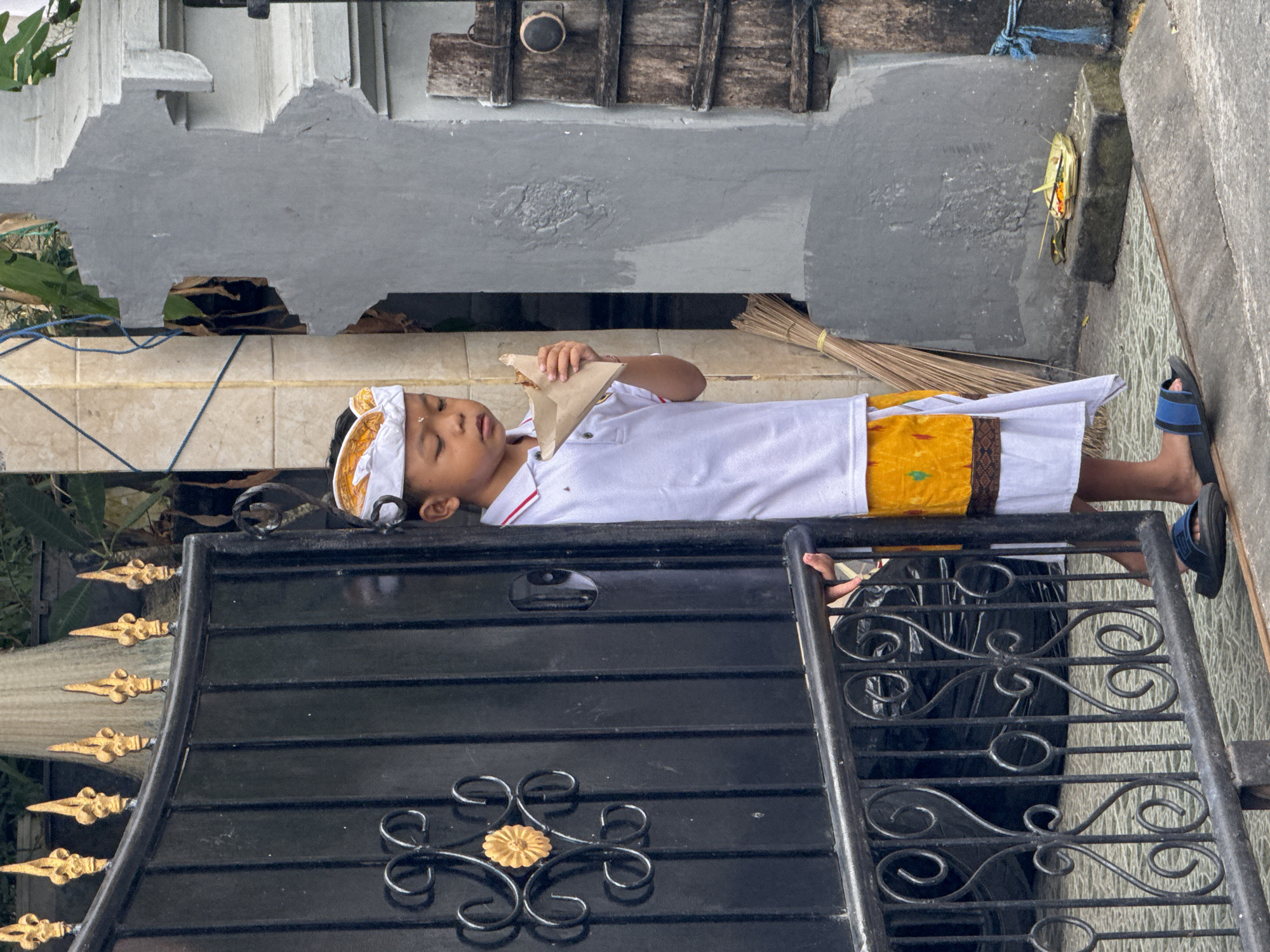 A young boy stands partially obscured by a black metal gate, holding a small, folded piece of brown paper containing food to his mouth. He is wearing a white polo shirt with a red collar, a yellow sash with an intricate pattern around his waist, and a white and gold headdress. His eyes are closed. The gate has ornate golden finials at the top and decorative scrollwork. Behind the boy, a textured grey wall with carved decorative elements and a wooden shutter can be seen. A broom leans against the wall. On the ground in front of the wall, a small offering made of leaves, flowers, and sticks rests on a stone block. The scene appears to be outdoors in an urban environment, possibly Kuta, Indonesia.