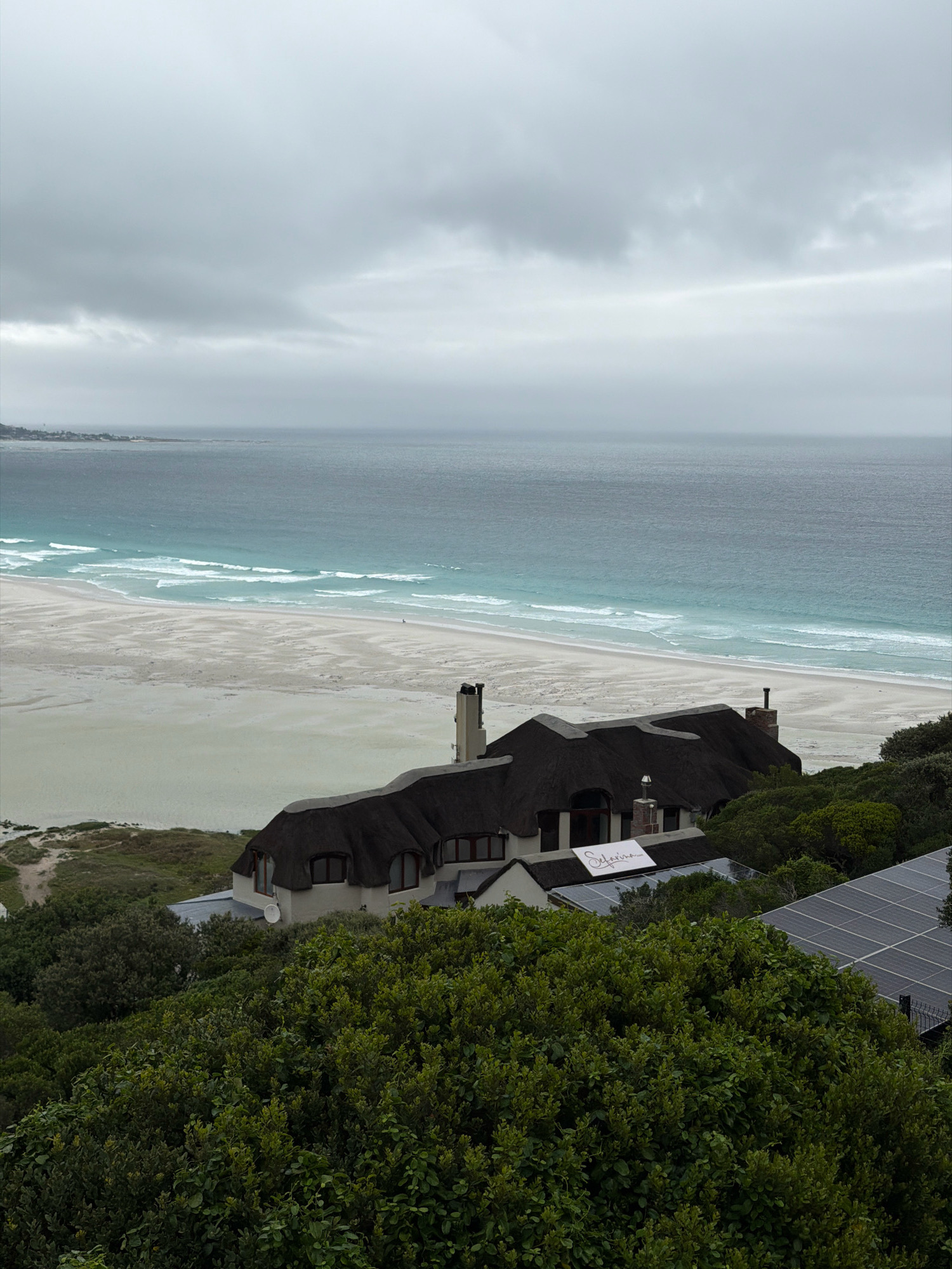A wide-angle, elevated shot captures a coastal view in Noordhoek, South Africa, on an overcast day. The scene is dominated by the expanse of the ocean, a pale turquoise where it meets a wide, sandy beach, and a darker, steely blue further out. Waves create white lines as they crash on the shore. A single, small human figure is visible on the beach. In the foreground, a large building with a thatched roof and chimneys stands on a hillside covered in dense green vegetation. A sign with the word "Safarina" is attached to the building. To the right of the building, several solar panels are visible, partially obscured by trees. The sky is heavy with grey clouds.